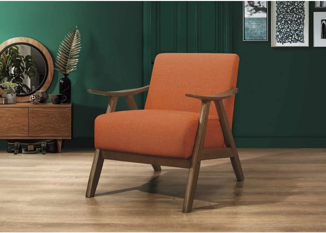 An orange upholstered armchair with wooden armrests and legs, placed on a light wood floor in a room with a dark green wall. To the left, there is a wooden sideboard with decorative items including a round mirror, a black vase with a large leaf, and small plants.