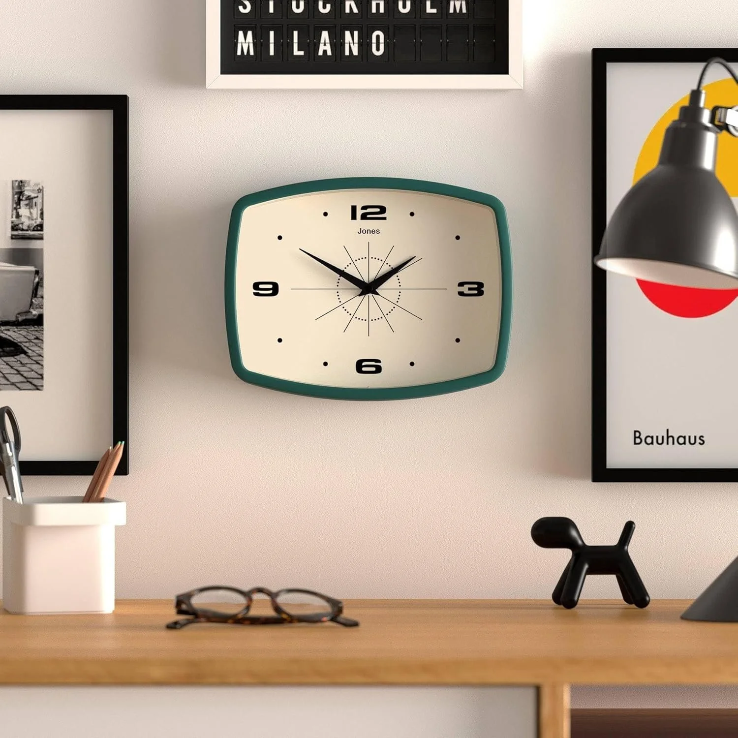 An MCM Jones wall clock with a green frame showing the time as 10:10 hanging on a white wall, surrounded by framed photos, a black dog figurine, a hanging lamp, and a small container with pencils on a wooden desk.
