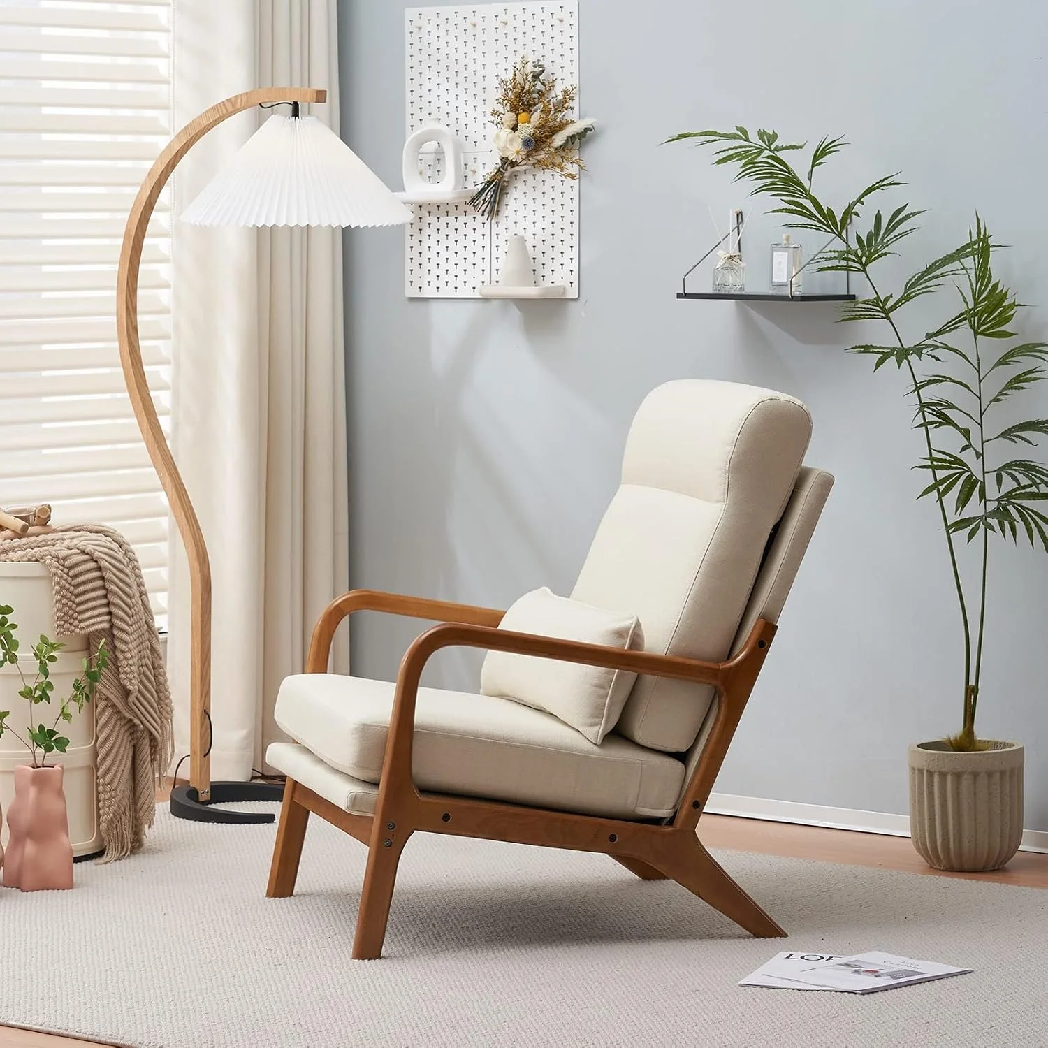 A cozy corner in a modern living space with a beige armchair with wooden arms, a standing lamp with a pleated white shade, a white pegboard wall with decorative items and with a bouquet of dried flowers, a black floating shelf with small plants and bottles, a large green plant in a beige pot, a small pink ceramic vase, and a magazine on a cream-colored rug.