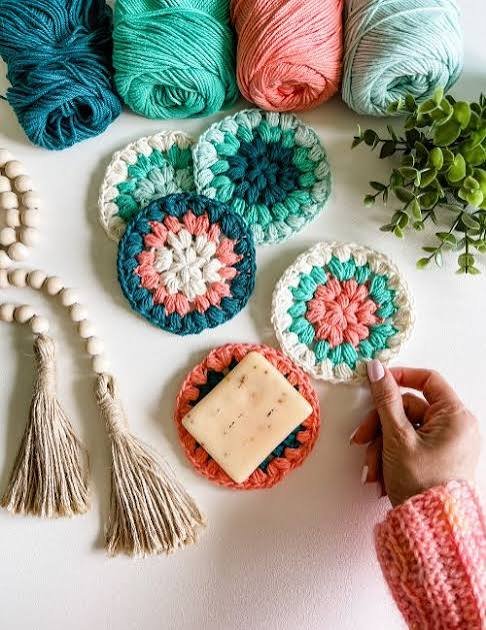 Various colorful yarn skeins, crocheted coasters, a bar of soap on a crocheted holder, and decorative beads and tassels on a white surface.