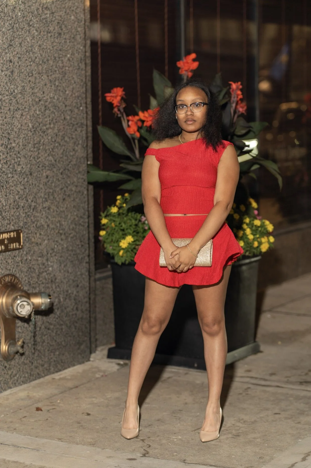 A young woman in a red off-shoulder dress stands in front of a flower arrangement at night, holding a clutch purse, wearing glasses and beige heels.