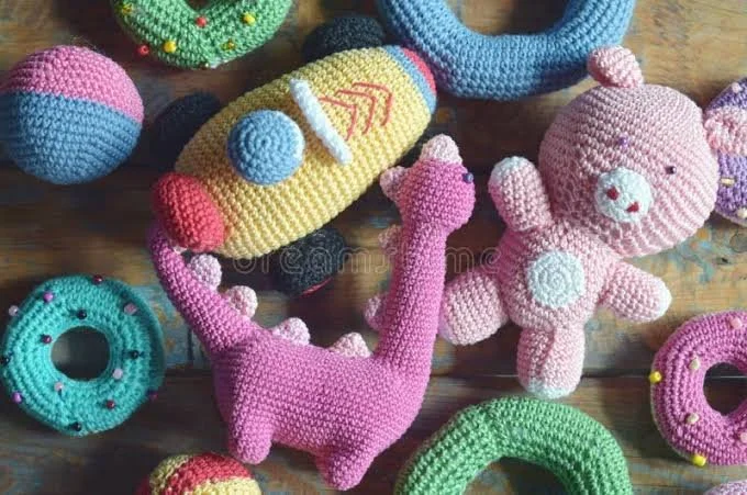 Colorful knitted plush toys including a pink dinosaur, a pink teddy bear, and various donut-shaped objects on a wooden surface.