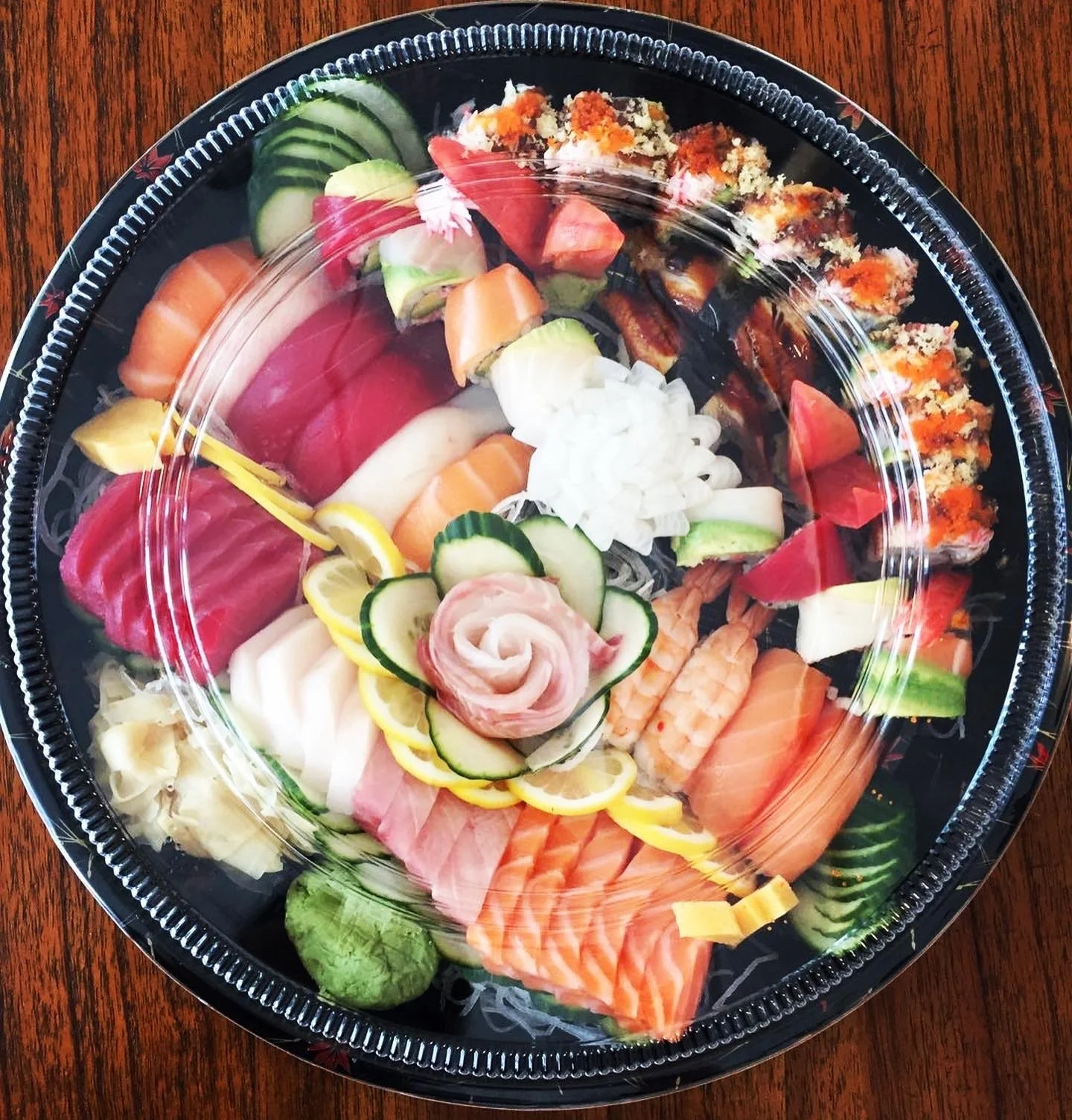 Assorted sushi platter with various types of fish, cucumber, avocado, lemon slices, and garnishes, covered with clear plastic lid on wooden table.