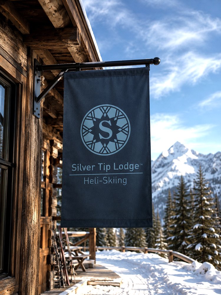 Blue sign hanging outside a wooden lodge with snow-covered mountains and trees in the background, indicating Silver Tip Lodge for helicopter skiing.