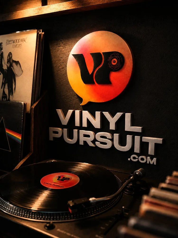 Vinyl record player with VinylPursuit.com sign on dark wall, also shows vinyl records on shelf.