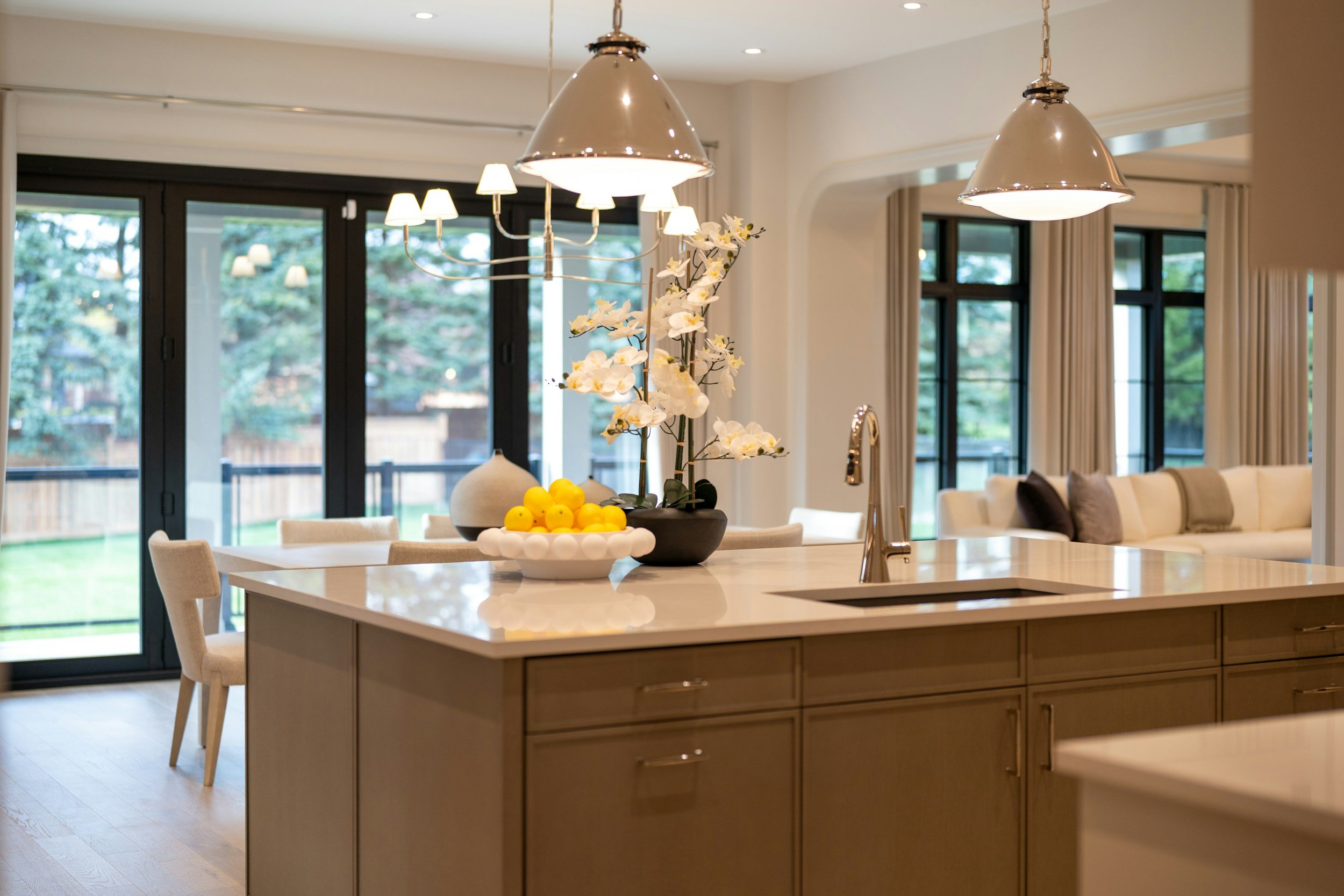 Modern kitchen with a white island, decorative flowers, lemons, and hanging pendant lights, with large windows and sliding glass doors in the background.