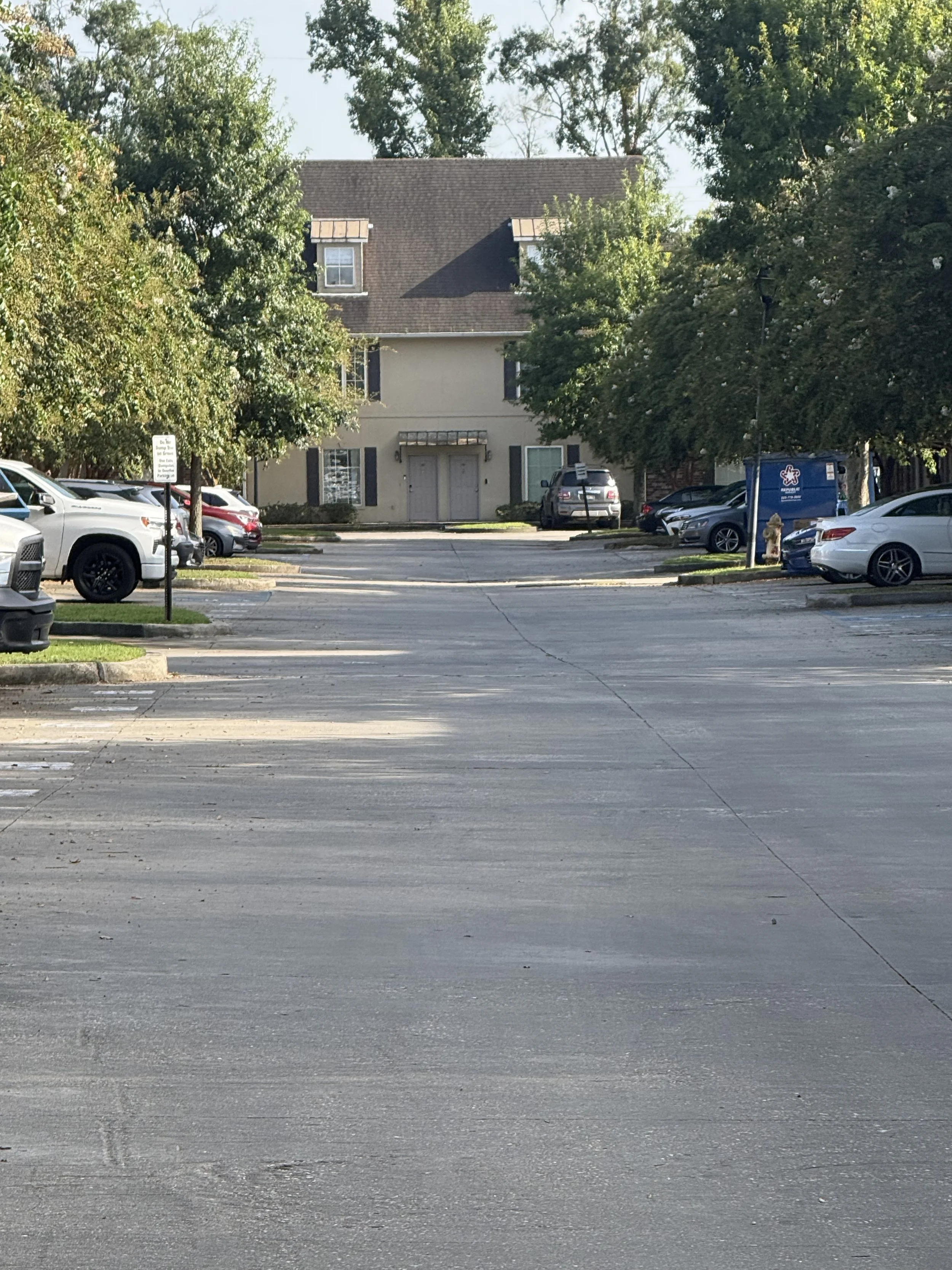 A parking lot with several parked cars in front of a beige apartment building with dark shutters and a brown roof, surrounded by trees.