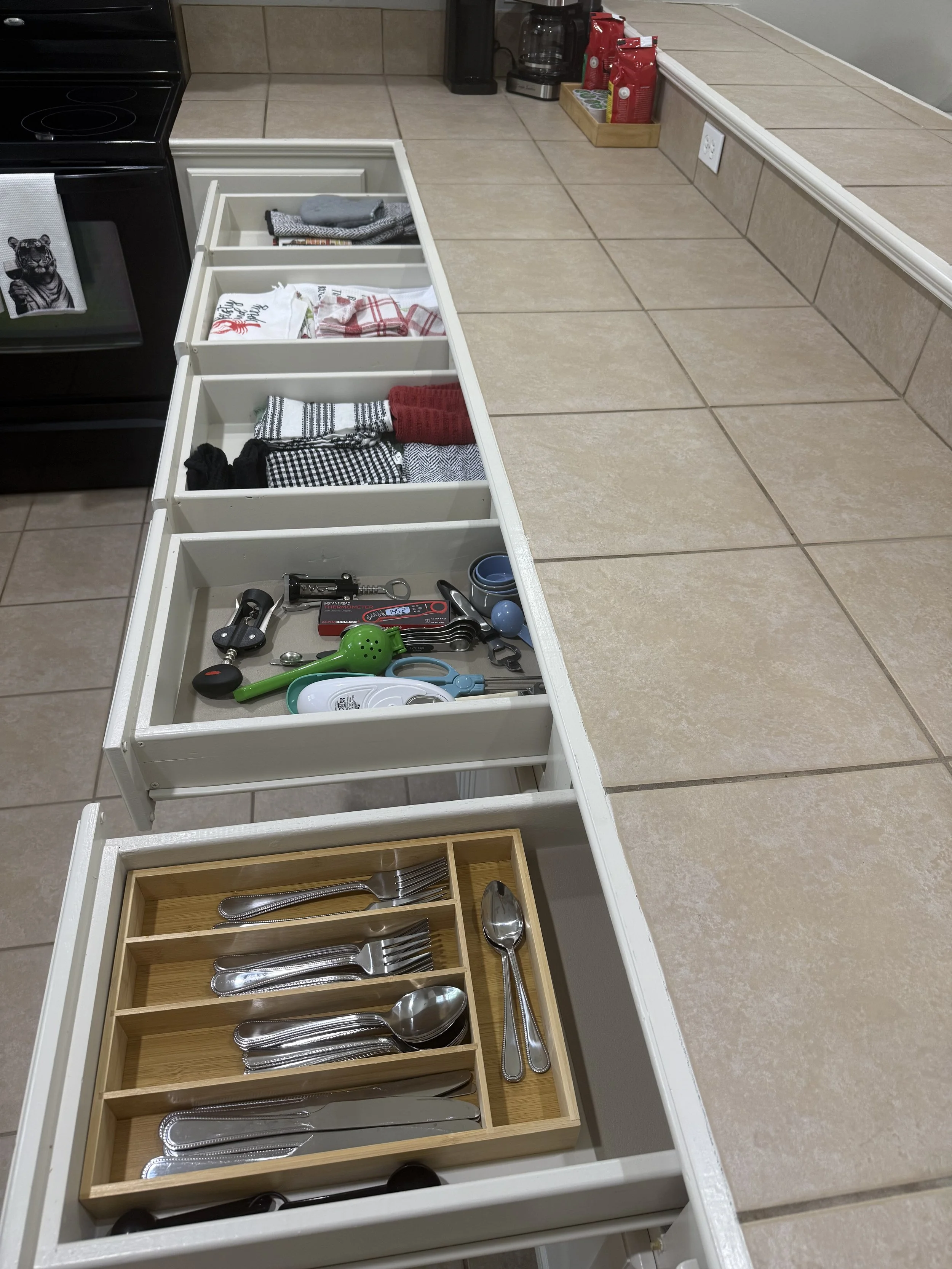 Kitchen counter with a white tray holding neatly folded tea towels, a gray cloth, and a black and white dish towel. Below the tray is a wooden cutlery organizer with silverware. The surrounding countertop has a coffee maker, coffee bags, a tray with 