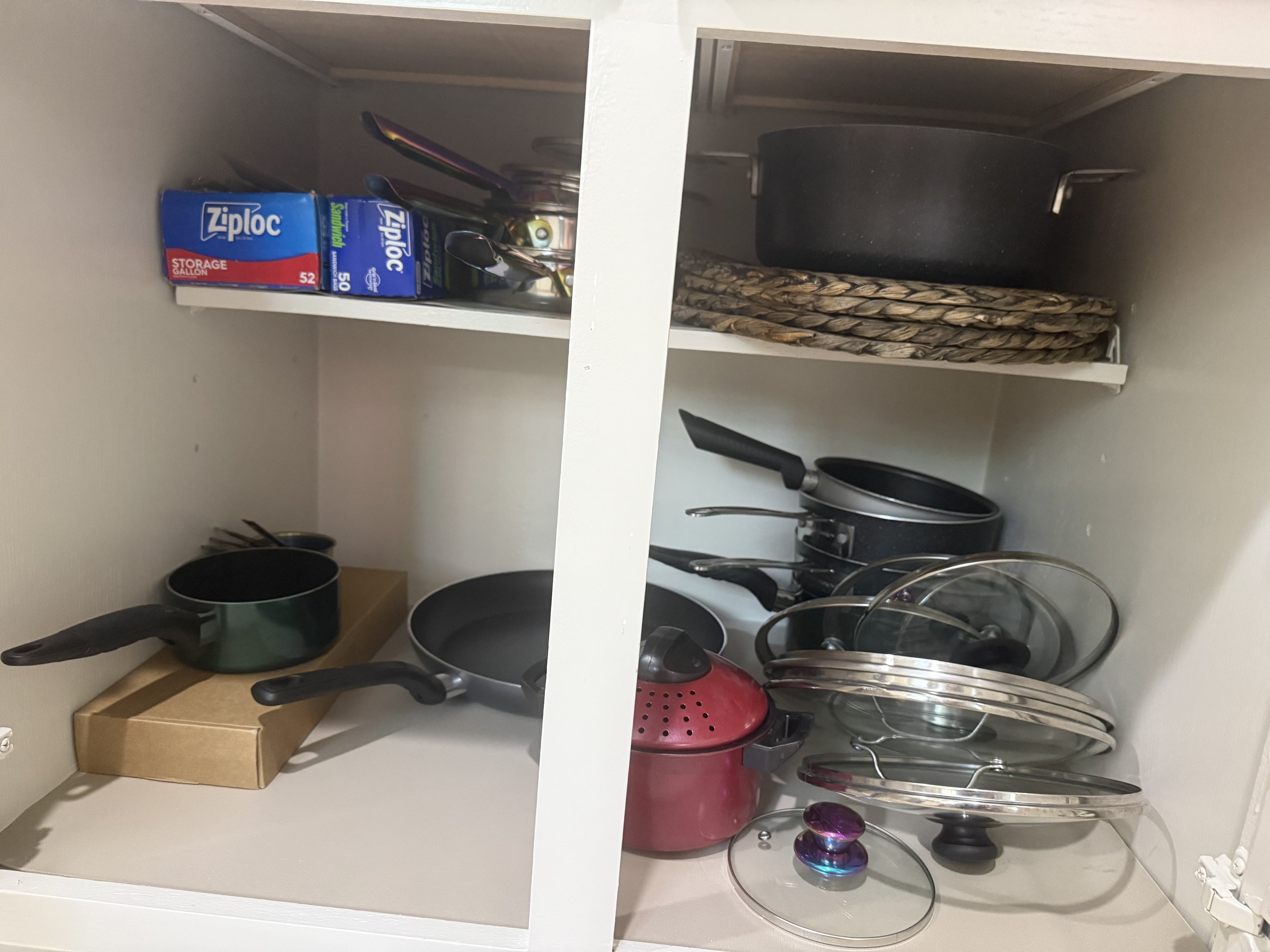 A cupboard with kitchen utensils and food storage items. The top shelf contains two boxes of Ziploc storage bags and some utensils. The bottom shelf has a variety of pots, pans, and lids, with some items stacked and some hanging on hooks.