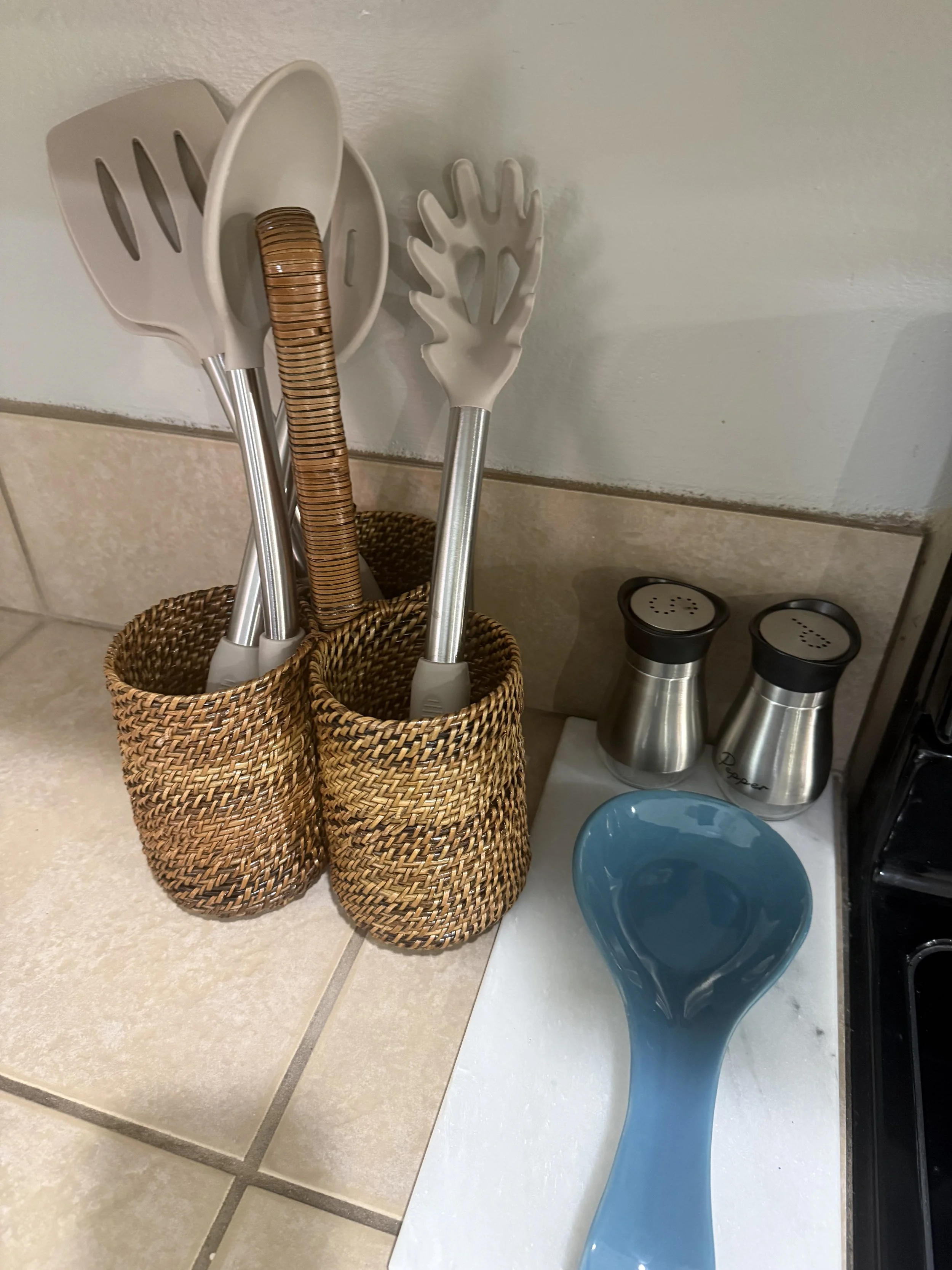 Kitchen countertop with two woven baskets holding kitchen utensils, including a spatula and a spoon, next to a pair of salt and pepper shakers and a blue glass serving dish.