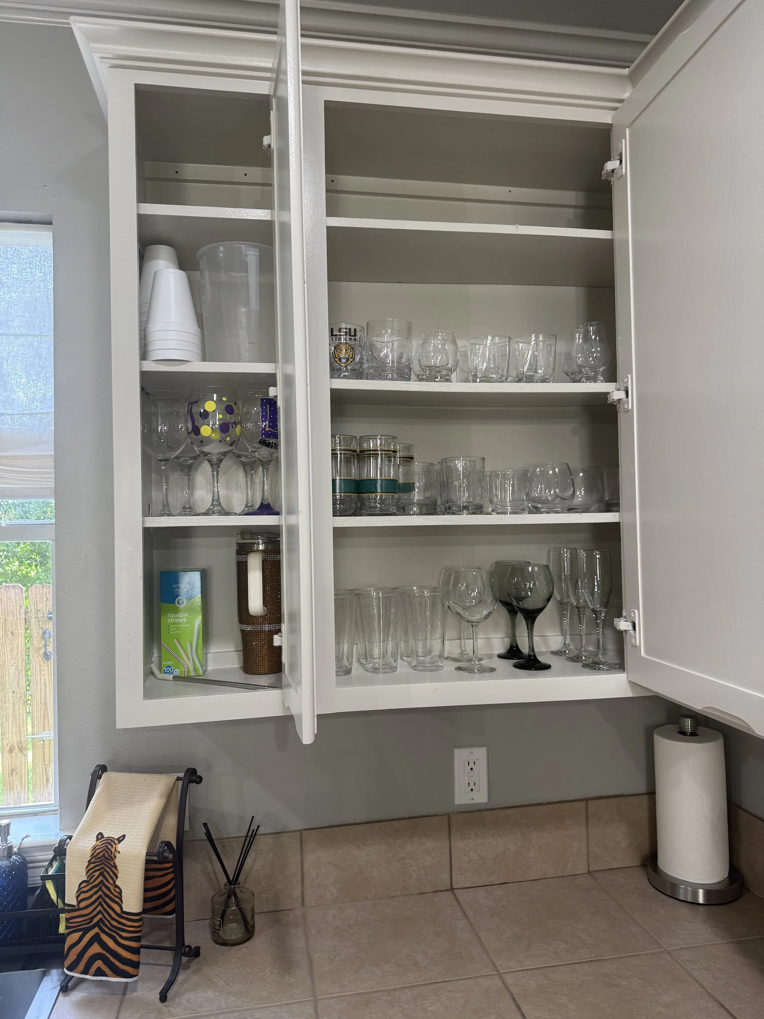 Open white kitchen cabinet with glassware on three shelves, including wine glasses, tumblers, and shot glasses. Items on the left include a plastic pitcher, paper cups, a box of straws, and a copper tumbler. There is a paper towel roll and a black ho