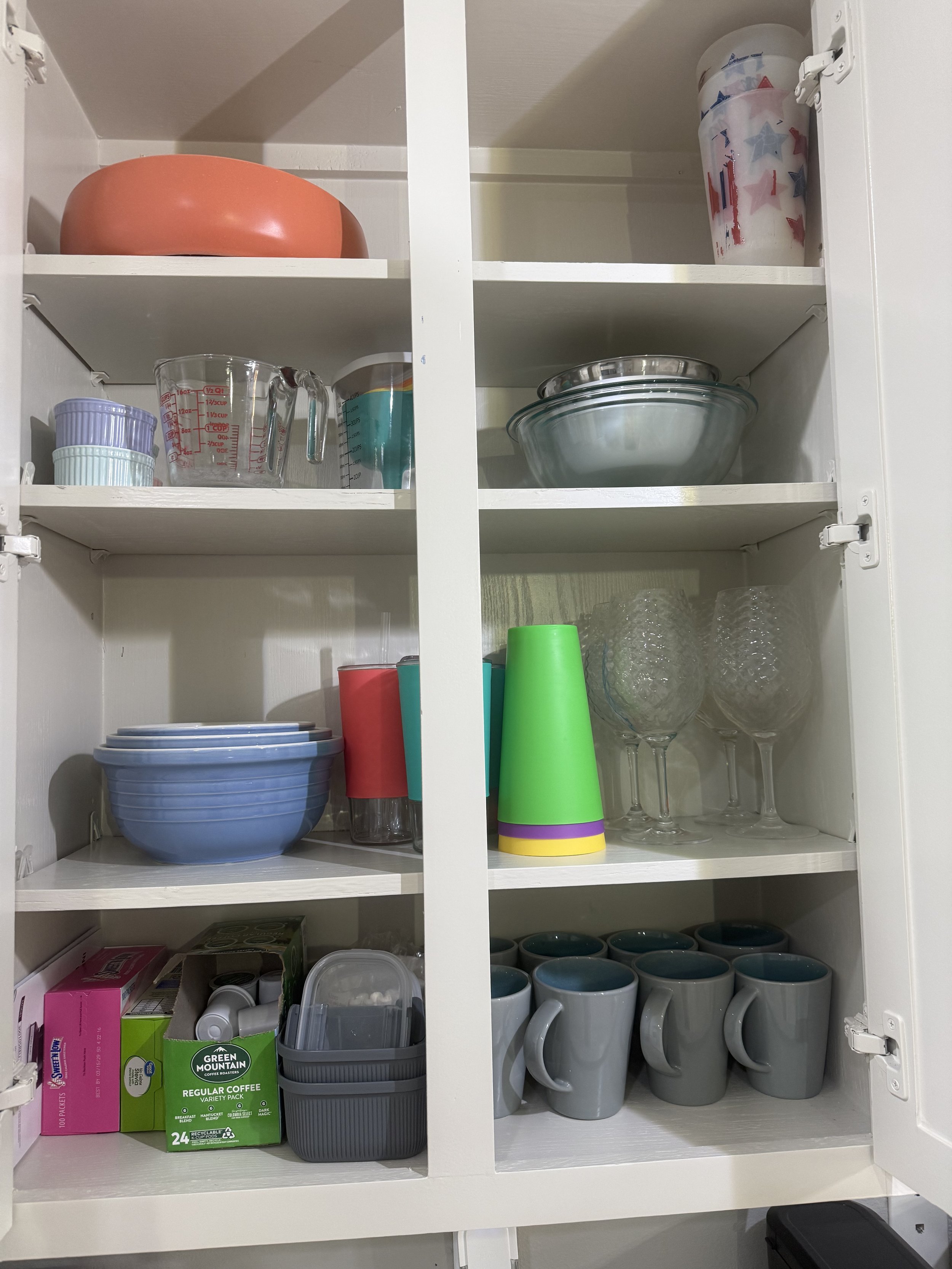 Kitchen cabinet filled with cups, bowls, glasses, mugs, cups, a blender, plastic containers, and boxes of coffee.