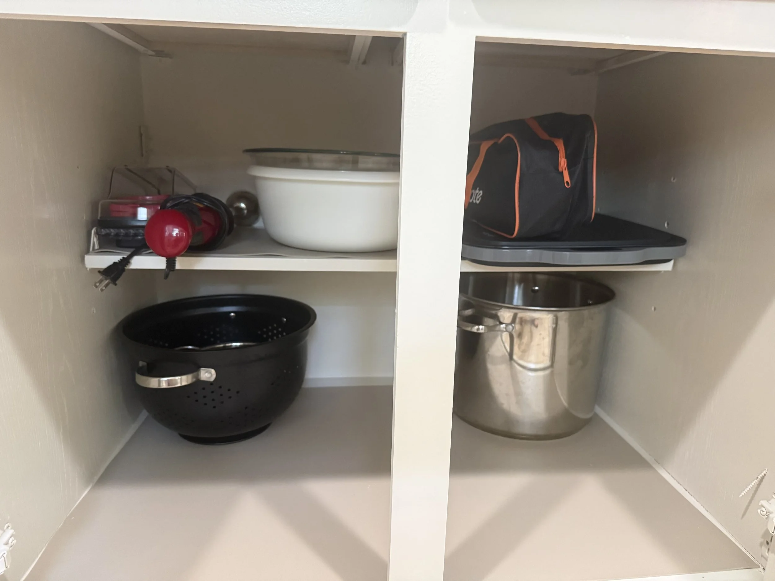 Inside a kitchen cabinet with two shelves holding various small appliances and kitchen items, including a black slow cooker, a colander, small containers, a black and orange tool bag, and a red handheld vacuum.