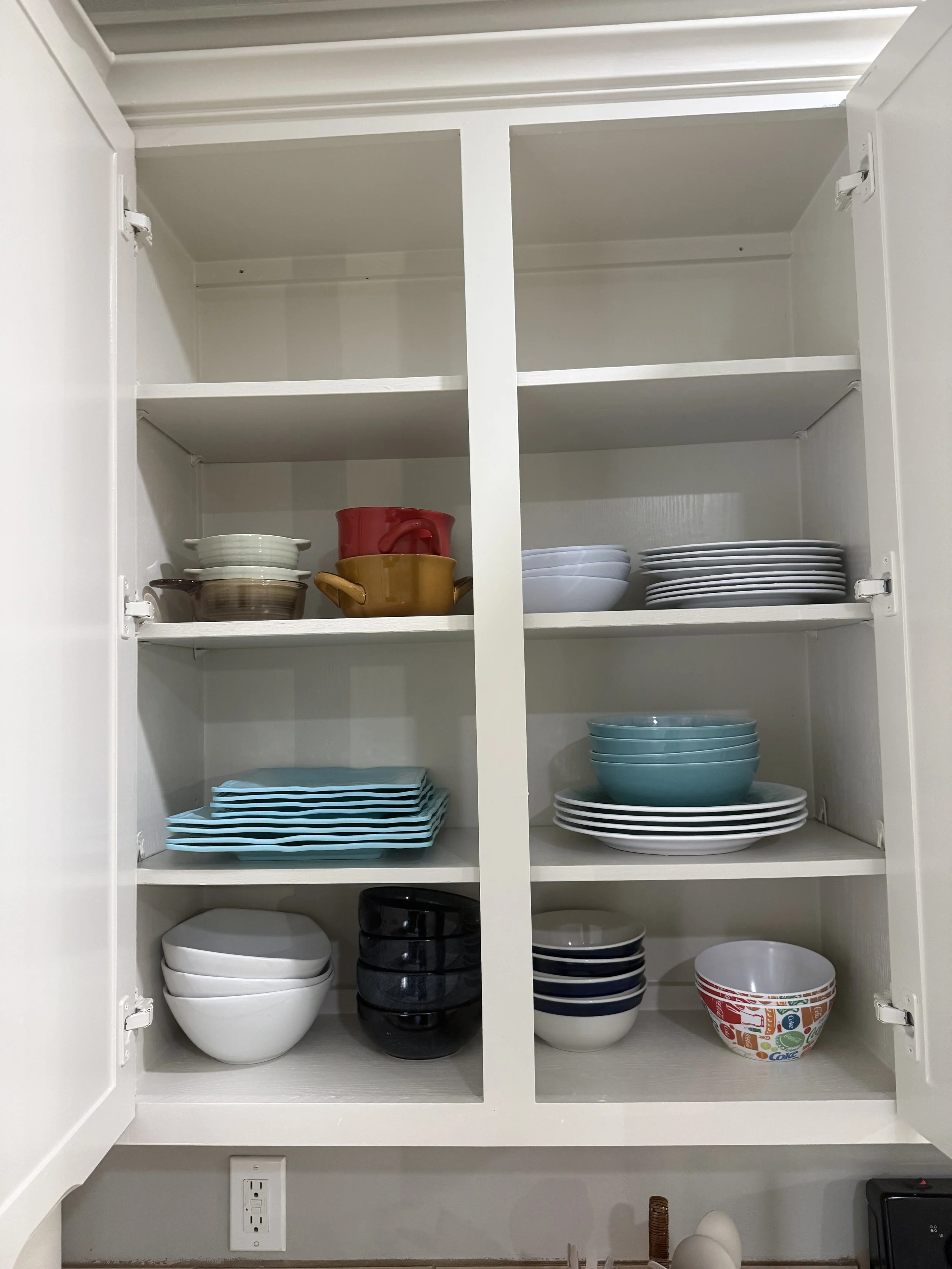 Open kitchen cabinet with plates, bowls, coffee mugs, and a Tupperware container inside.
