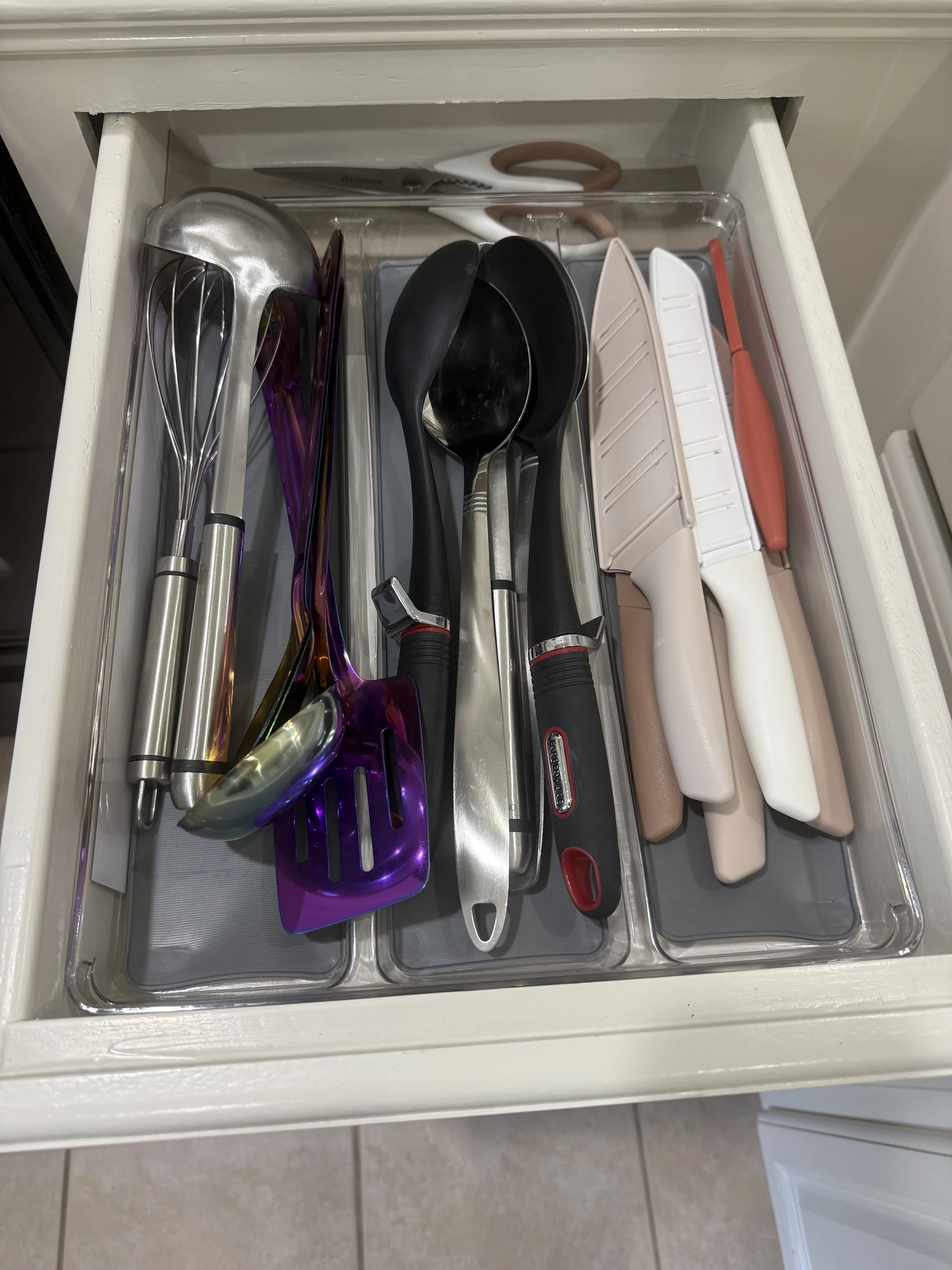 Drawers containing kitchen utensils, including a slotted spoon, ladle, spatula, and tongs, with a pair of scissors placed on top.