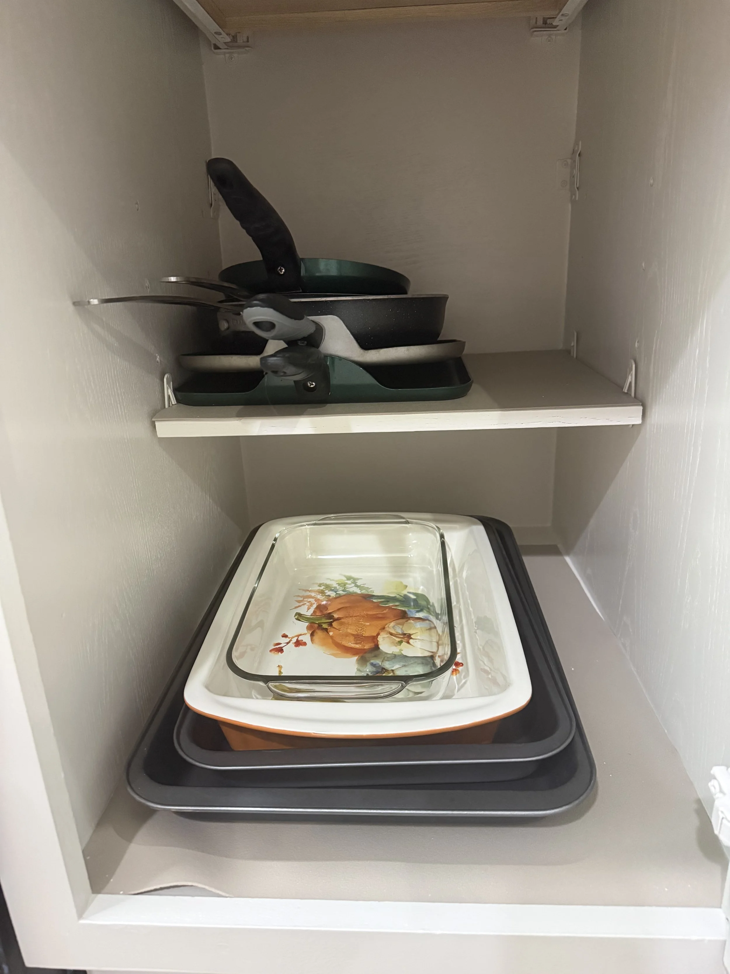 Inside a cabinet with a white trays containing a decorative glass dish with a pumpkin design, and two pans on a shelf above.