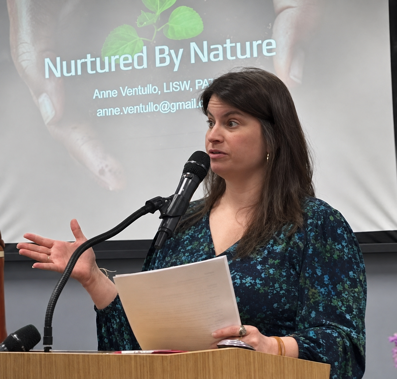 A woman standing at a podium giving a presentation, with a slide behind her titled 'Nurtured By Nature' showing her name, credentials, and email address. She is holding papers, speaking into a microphone, and gesturing with her other hand.