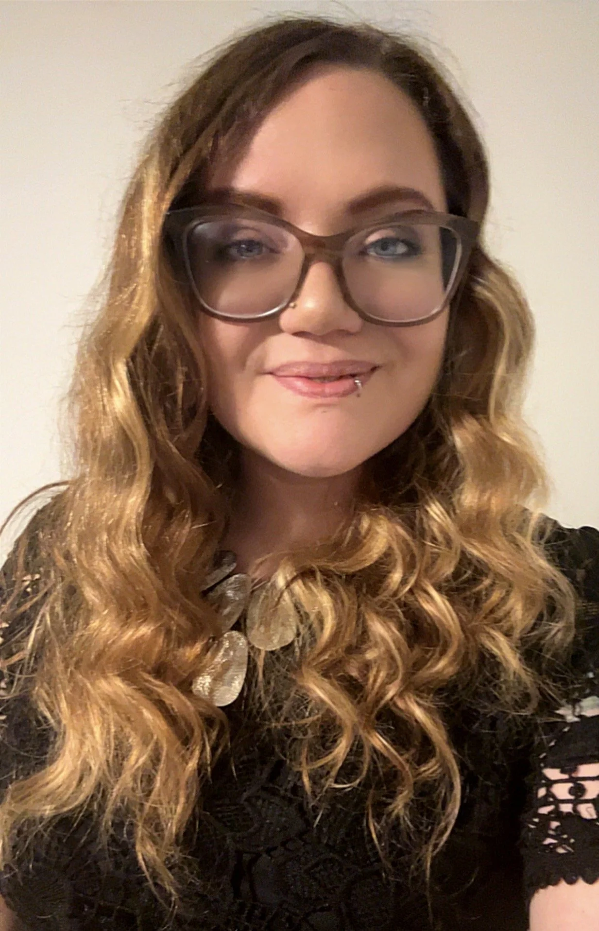 A woman with long, curly, ombré hair (dark brown to blonde) wearing glasses with dark frames, a black lace top, and makeup with pink lipstick. She is smiling slightly and sticking out her tongue with a piercings on her lower lip.