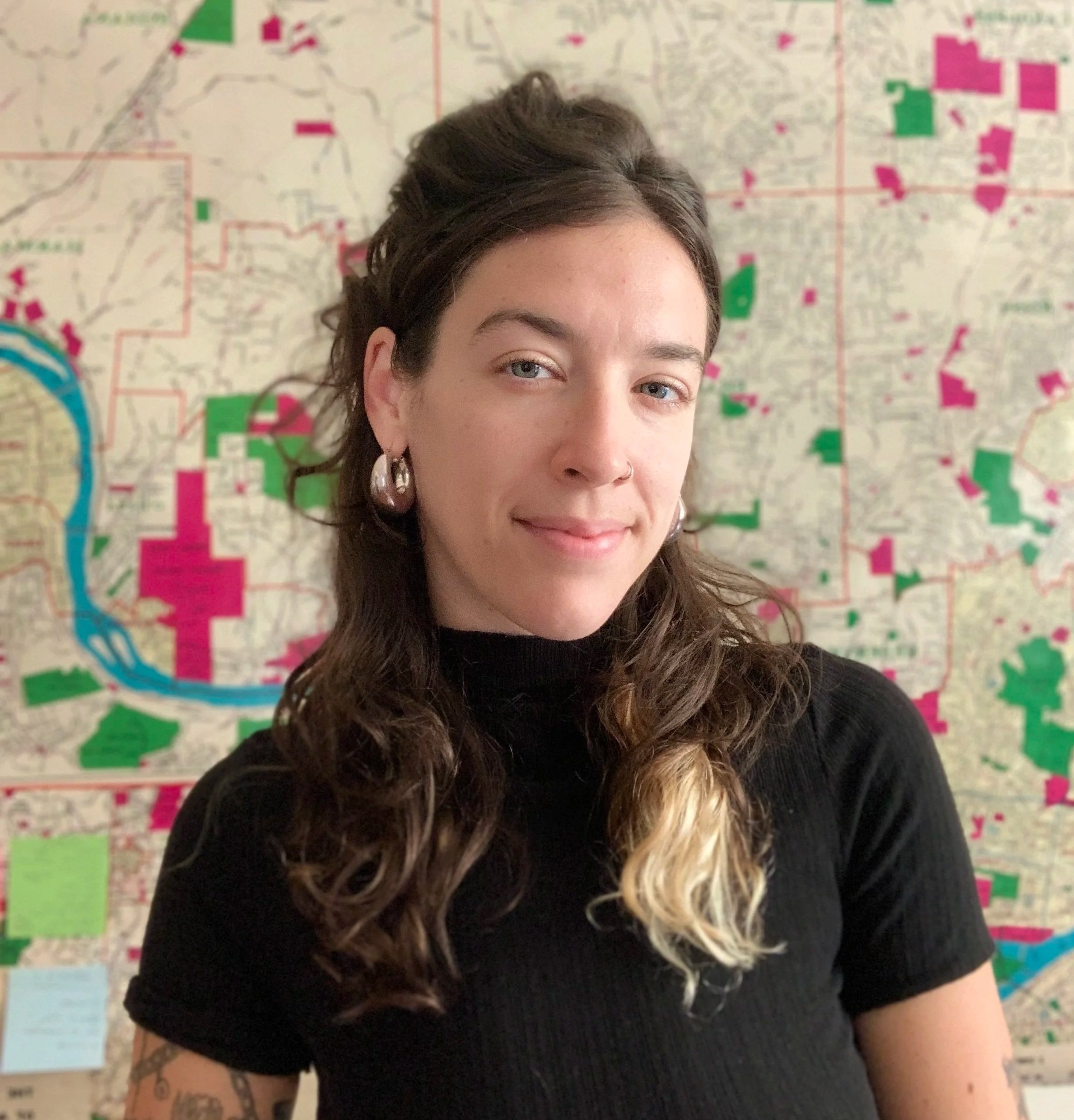 Close-up of a person with dark brown and blonde curly hair, wearing a black shirt and earrings, standing in front of a colorful map on the wall.