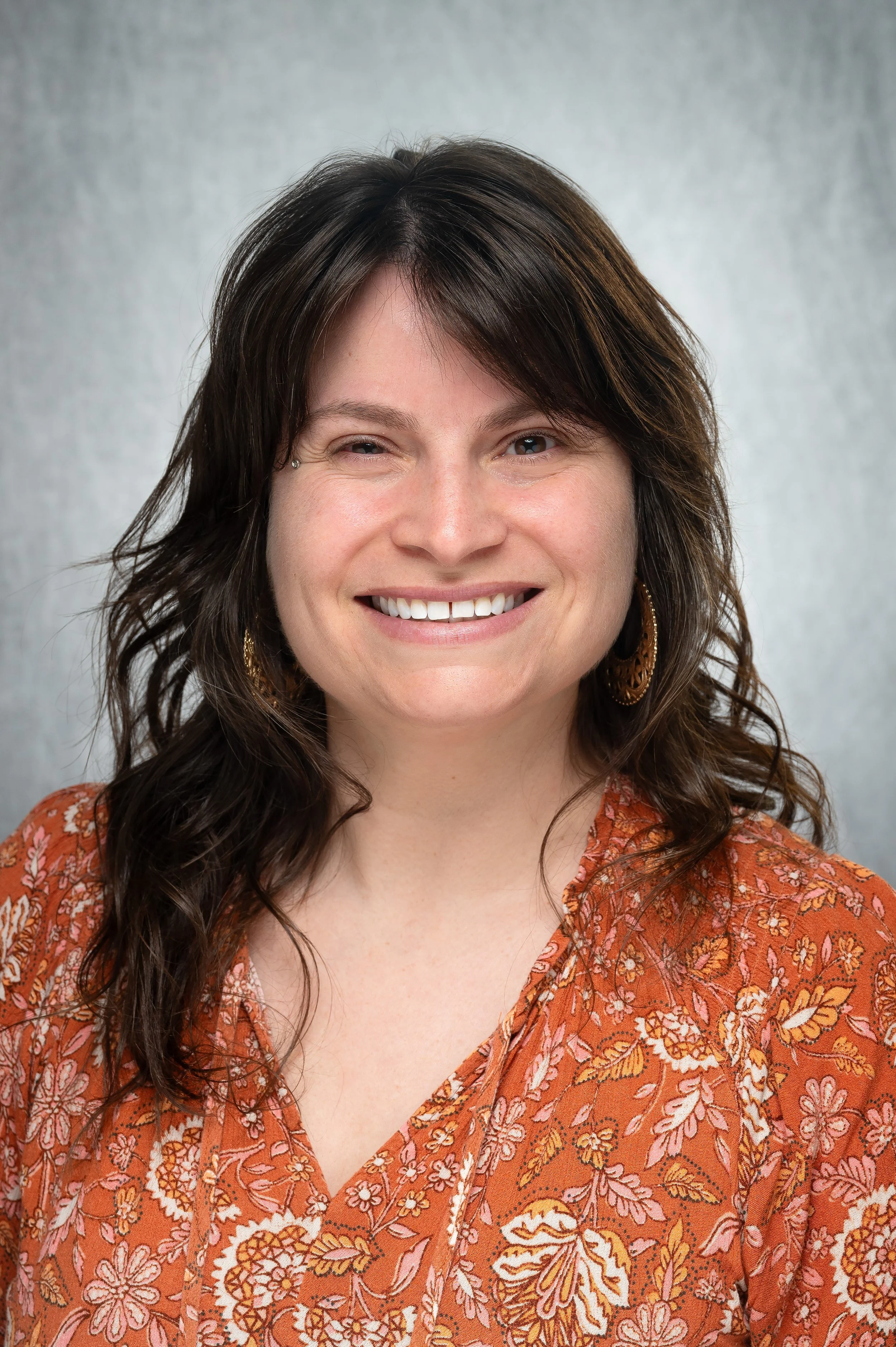 Close-up of a smiling woman with long, wavy dark hair wearing a burnt orange floral blouse and gold earrings, standing against a plain grey background.