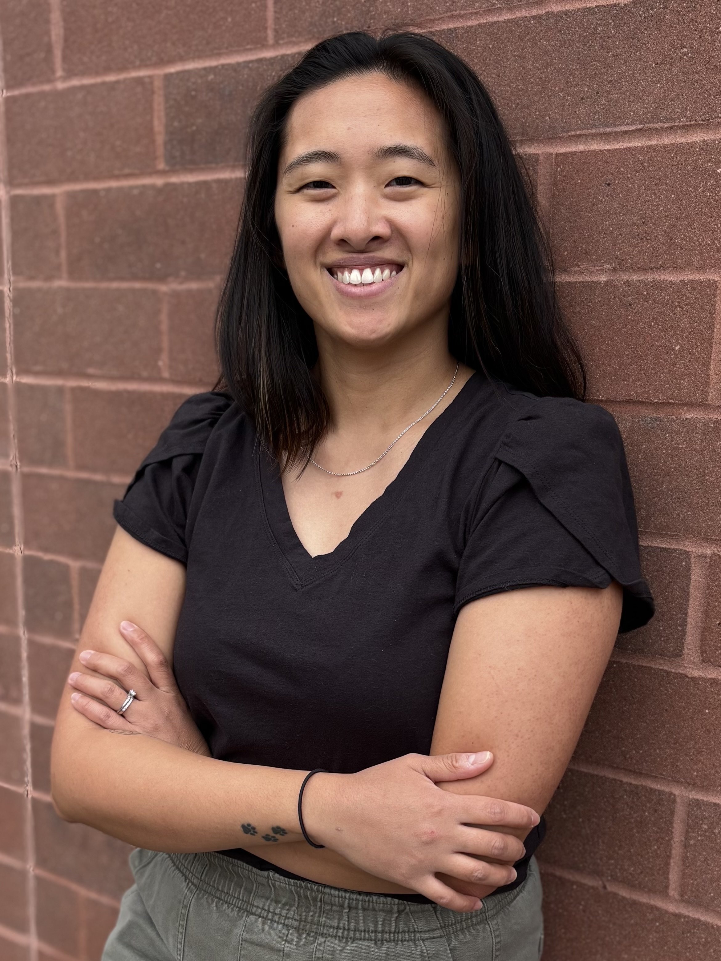 A woman with long black hair, smiling, standing against a brick wall. She is wearing a black shirt and gray pants, with her arms crossed. She has a small tattoo of holly leaves on her left wrist, a ring on her left hand, and a necklace.