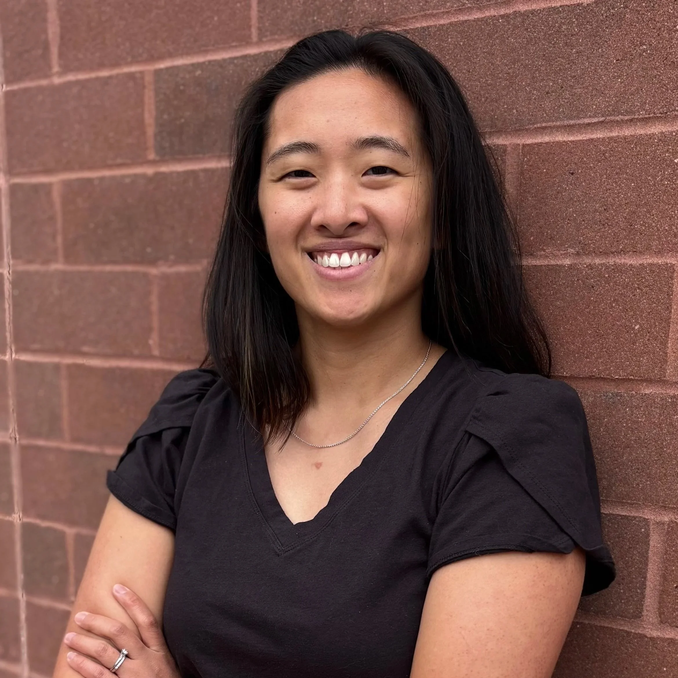 Close-up of a smiling woman with long dark hair, wearing a black top, standing against a brick wall.