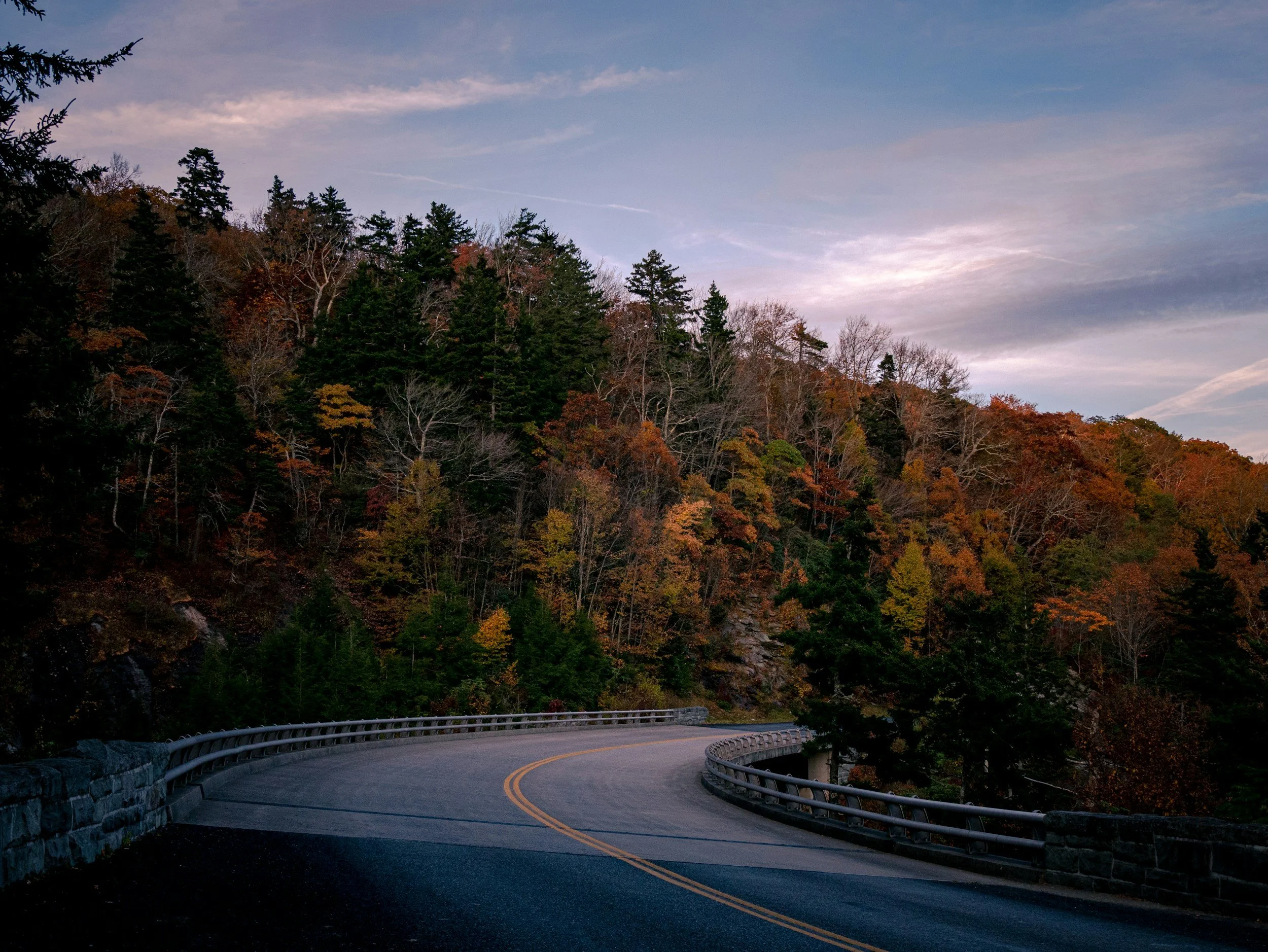 A winding mountain road with a guardrail on the side, surrounded by dense trees with autumn foliage, under a partly cloudy sky.