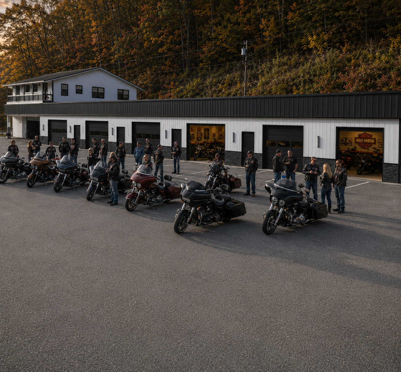 A group of people standing near parked motorcycles in front of a building with garage doors, in a parking lot with trees and a wooden house in the background.