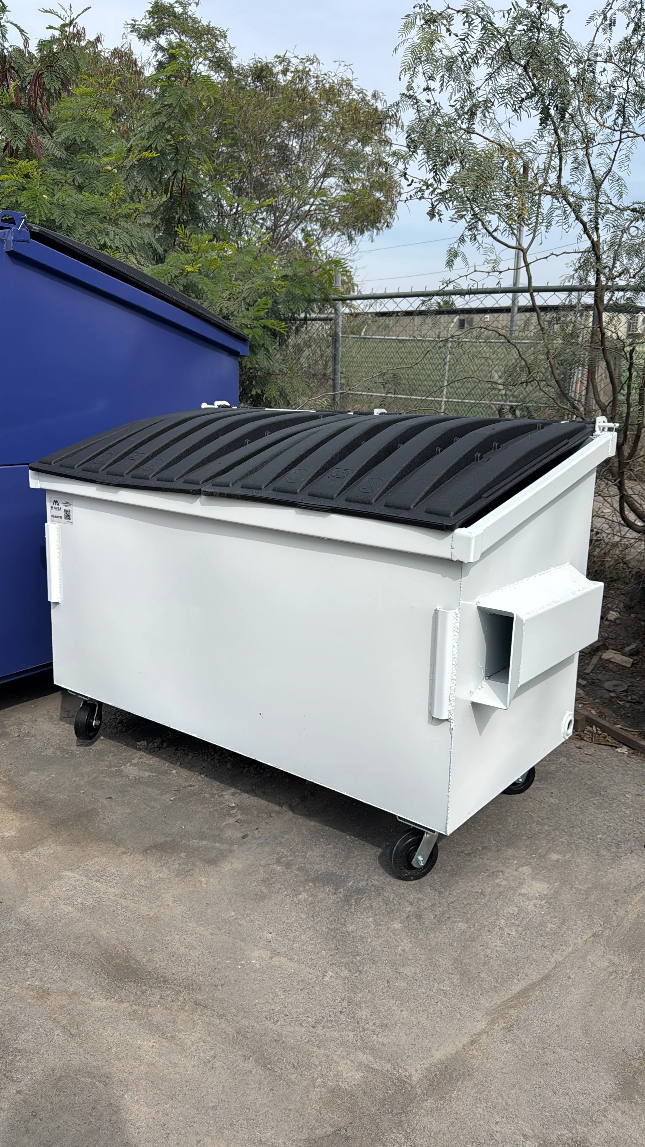 A white, wheeled dumpster with a black lid is outdoors on a concrete surface, next to a blue container and some trees and a chain-link fence in the background.
