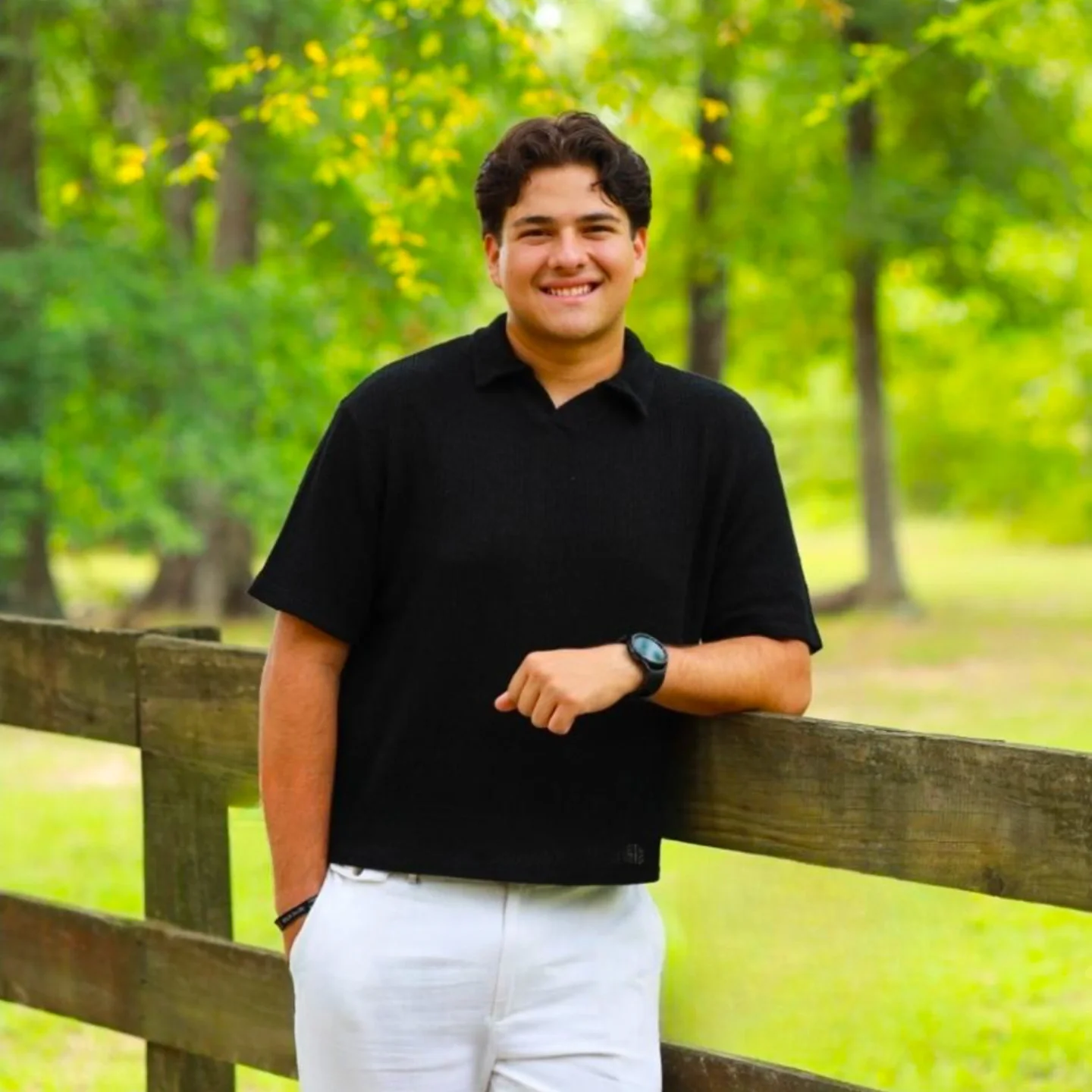 A young man with dark hair and a smile, wearing a black shirt and white pants, standing outdoors near a wooden fence in a green, wooded area.