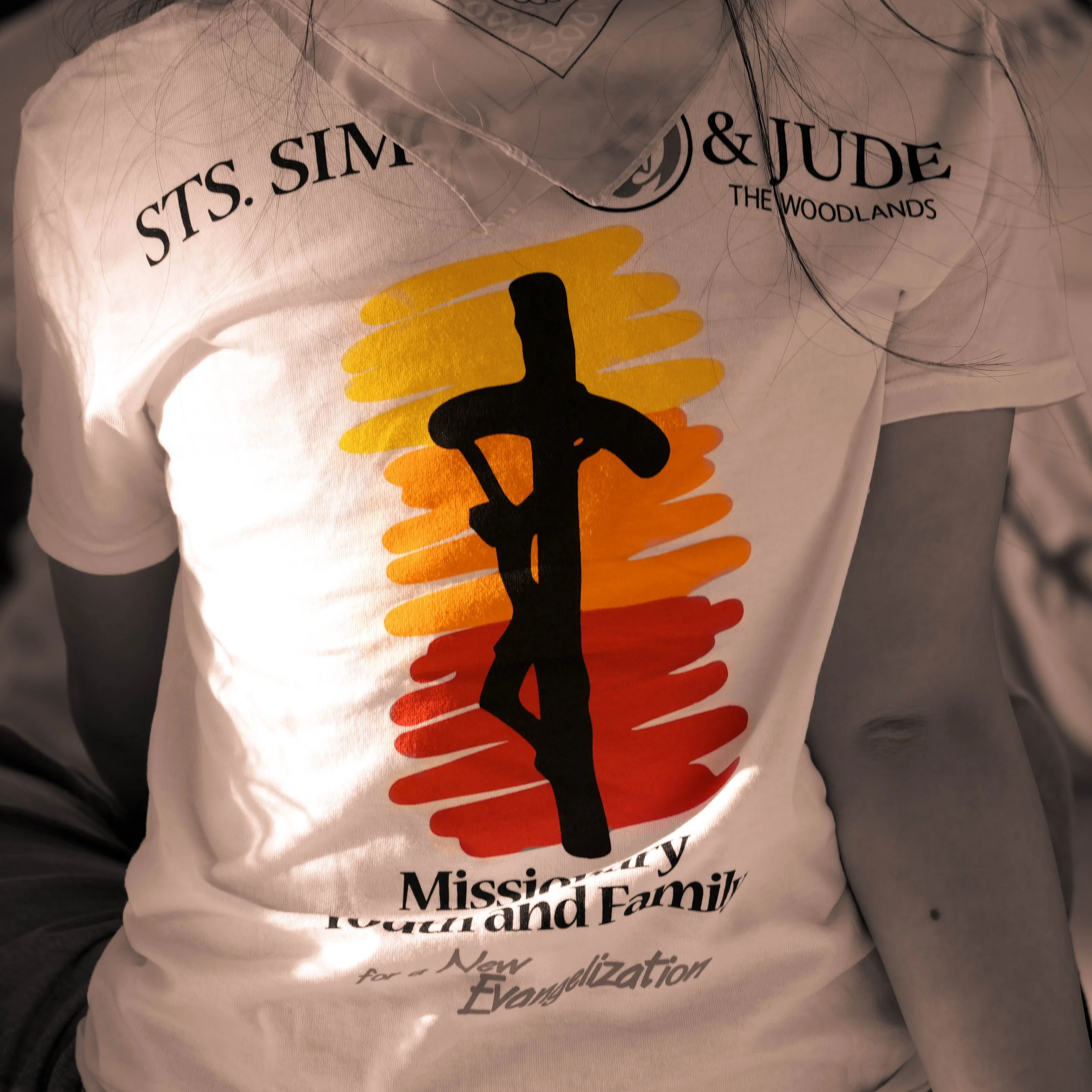 Close-up of a white T-shirt with black, yellow, and orange text and graphics. The shirt features a message supporting a family-based immigration reform for Muslim, Sikh, and Hindu communities in Massachusetts. The design includes a black silhouette of a person and the words 'Muslim Family', 'Massachusetts', and references to a new immigration reform.