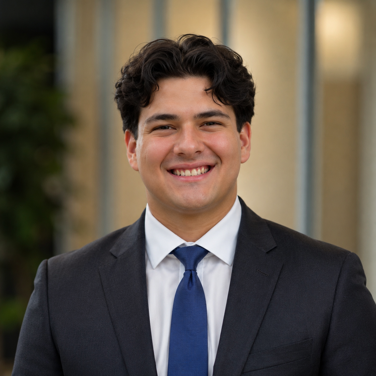 A young man in a dark suit, white shirt, and blue tie, smiling warmly outdoors near a building with windows and greenery.