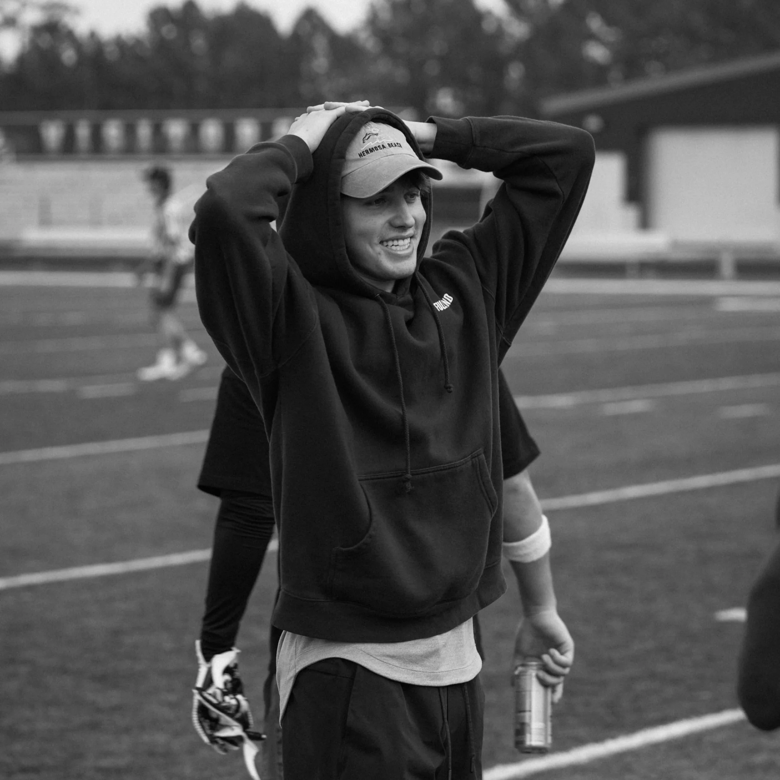 A young person wearing a hoodie and cap holding a drink bottle while standing on a running track, with other athletes blurred in the background.