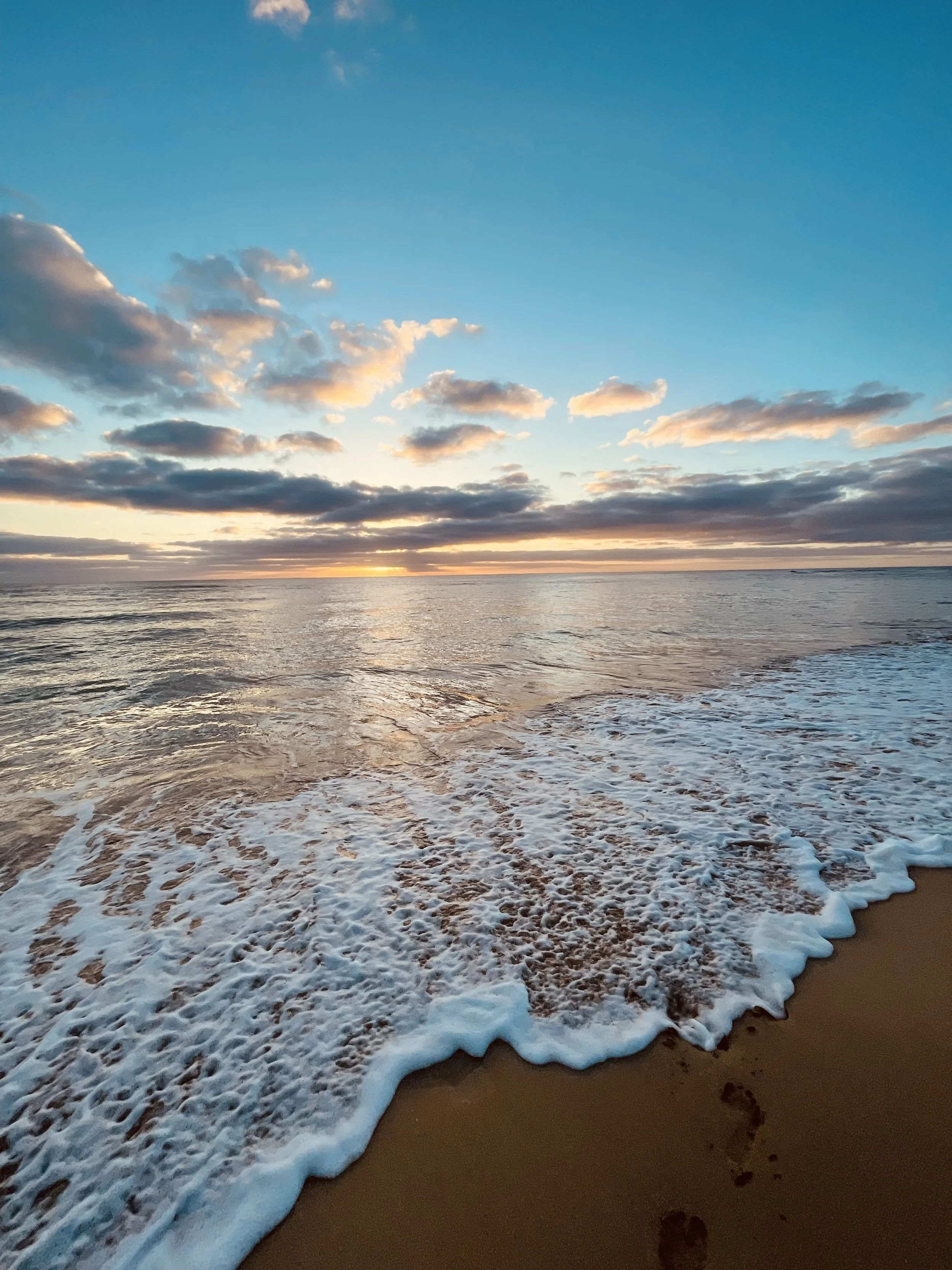Sunset over the ocean with clouds and waves crashing on the sandy beach, footprints in the sand.
