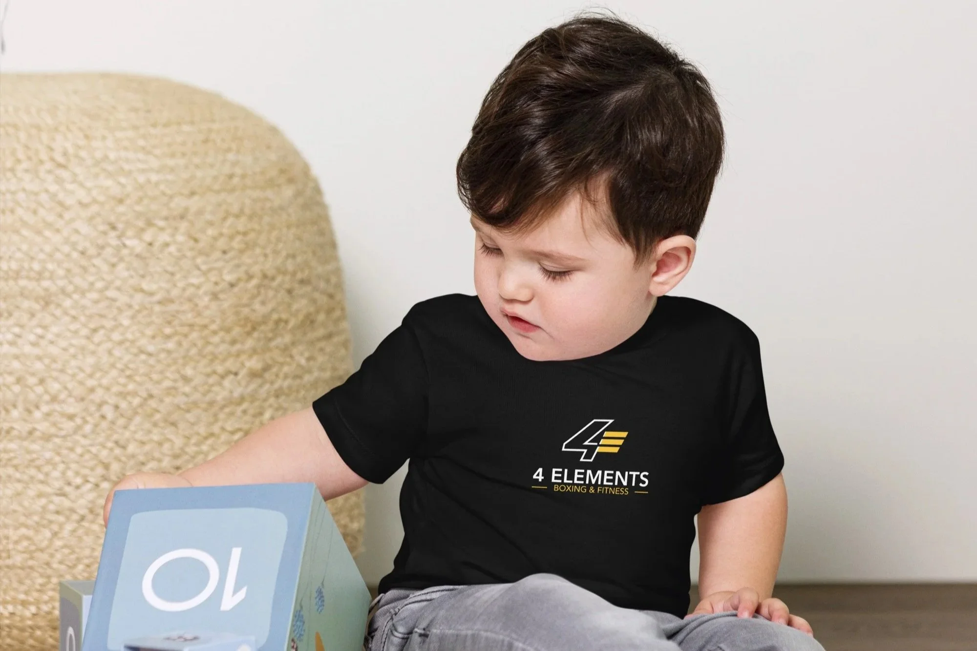 A young boy with dark brown hair, wearing a black t-shirt with the logo and text '4 ELEMENTS BOXING & FITNESS,' sitting on the floor and looking down at a blue foam block with letters on it, next to a beige textured object and some gray floor.