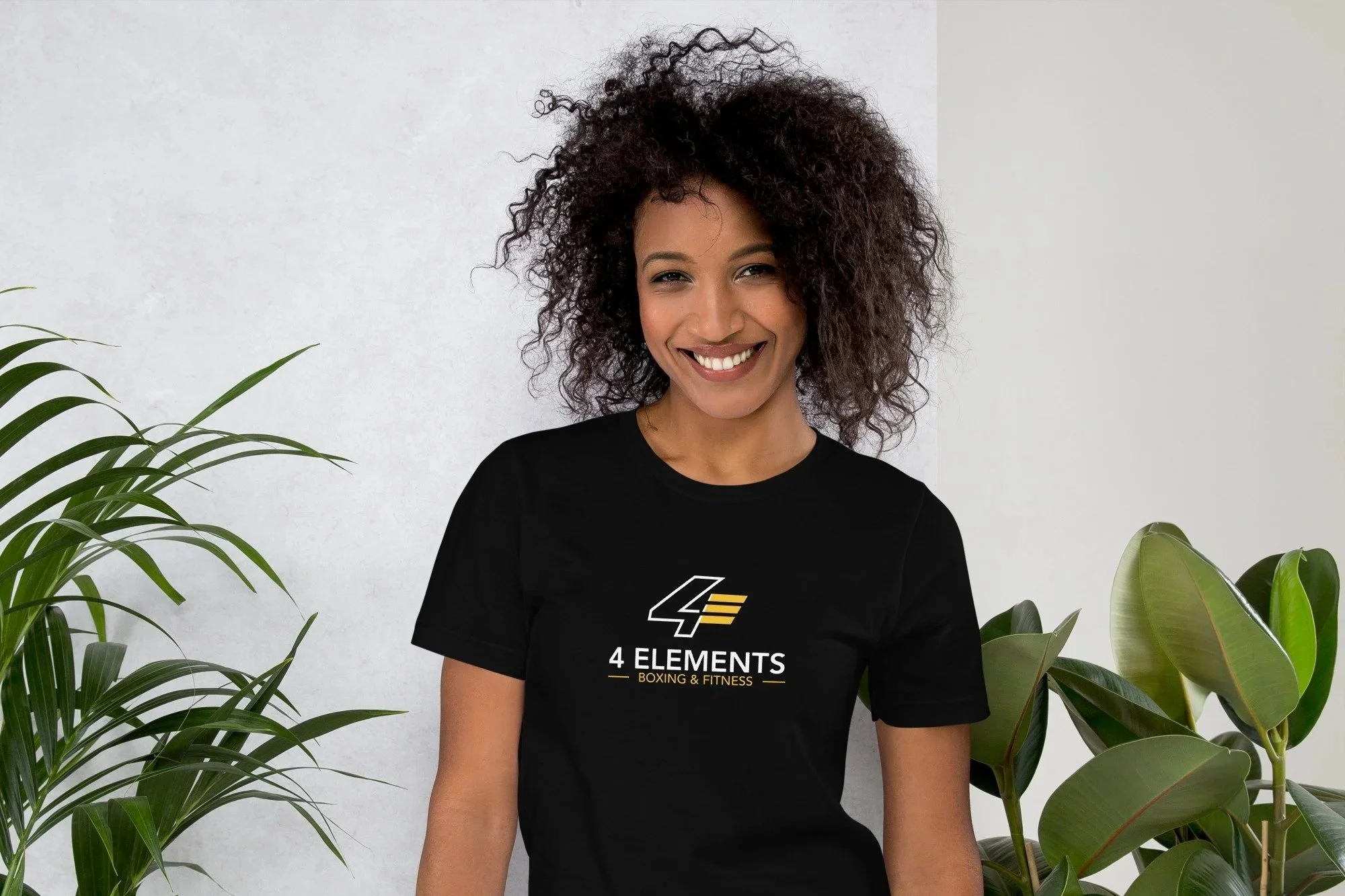 A woman with curly hair smiling, wearing a black t-shirt with a logo and text that says '4 ELEMENTS BOXING & FITNESS'. She is standing among green houseplants in front of a white wall.
