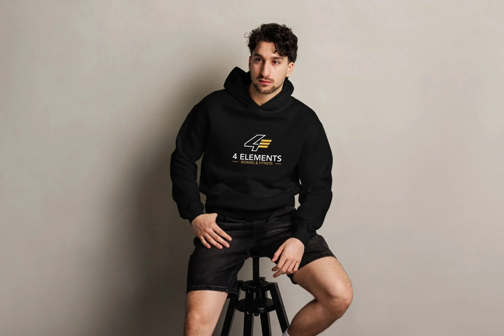 A young man with dark curly hair and a beard sitting on a black stool against a plain beige wall. He is wearing a black hoodie with the logo '4 Elements Boxing & Fitness' and black shorts. His right hand is resting on his thigh, and his left hand is in his pocket.
