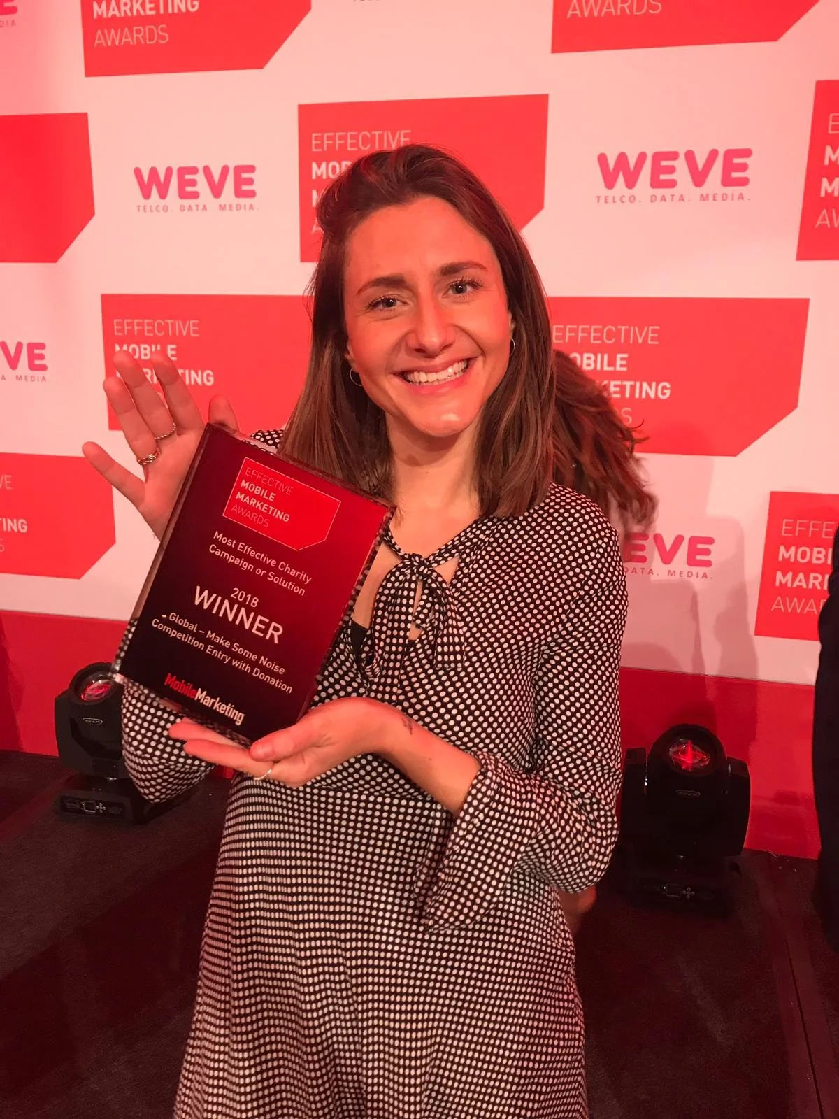 Woman smiling and holding an award plaque at a conference with a red and white backdrop.