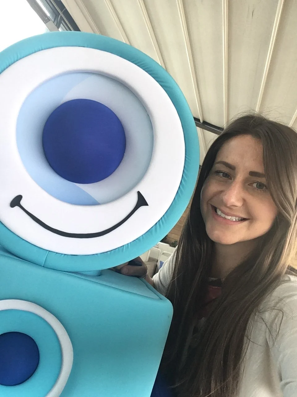A woman with long brown hair smiling next to a large blue cartoon character with a big eye and a smile, set against a ceiling with metal beams.