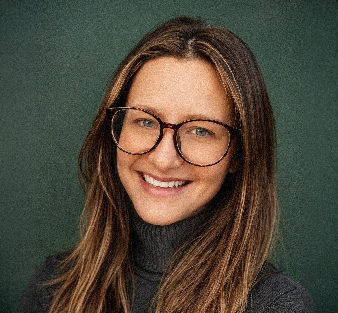 A smiling woman with long brown hair, glasses, and a dark turtleneck against a green background.