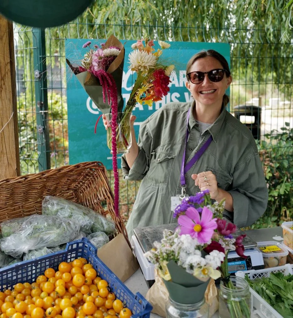 A woman smiling and wearing sunglasses, holding a bouquet of flowers at a market or stall, with various flowers, vegetables, and baskets on the table.