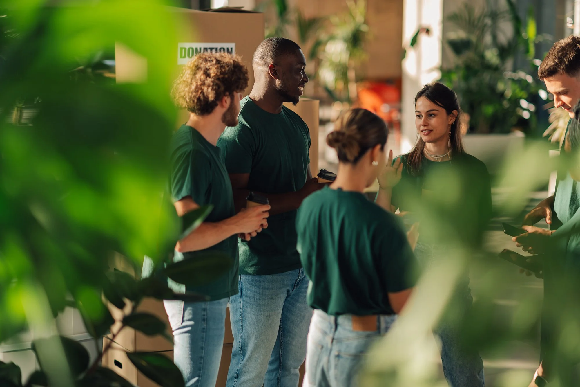 Group of diverse people at a donation event, standing indoors, holding coffee cups, engaging in conversation, with donation boxes in the background.