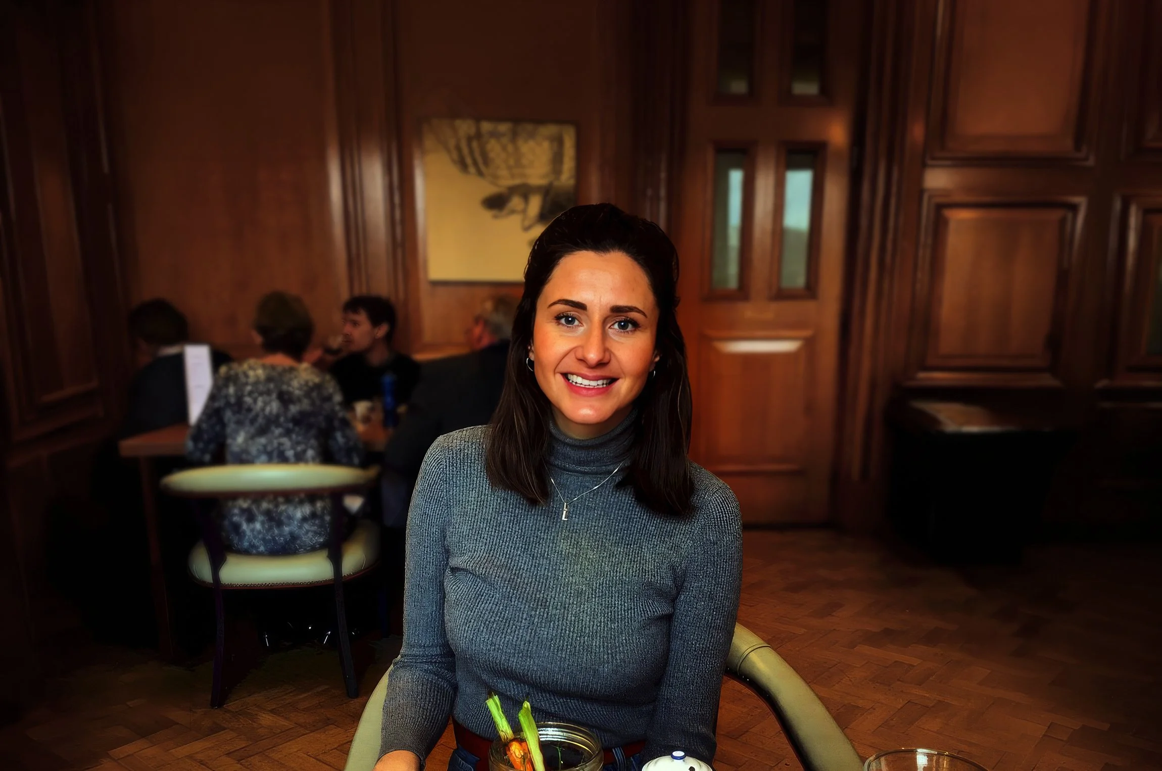 A woman with dark hair and blue eyes sitting at a table in a restaurant, smiling at the camera. She is wearing a gray turtleneck sweater, and there are other diners in the background with wooden paneling on the walls.