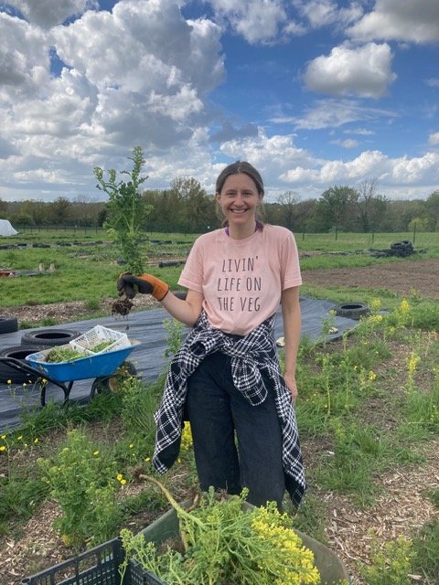 A woman in a pink T-shirt and dark pants holding a small plant in a garden or farm field, smiling at the camera with a partly cloudy sky overhead.