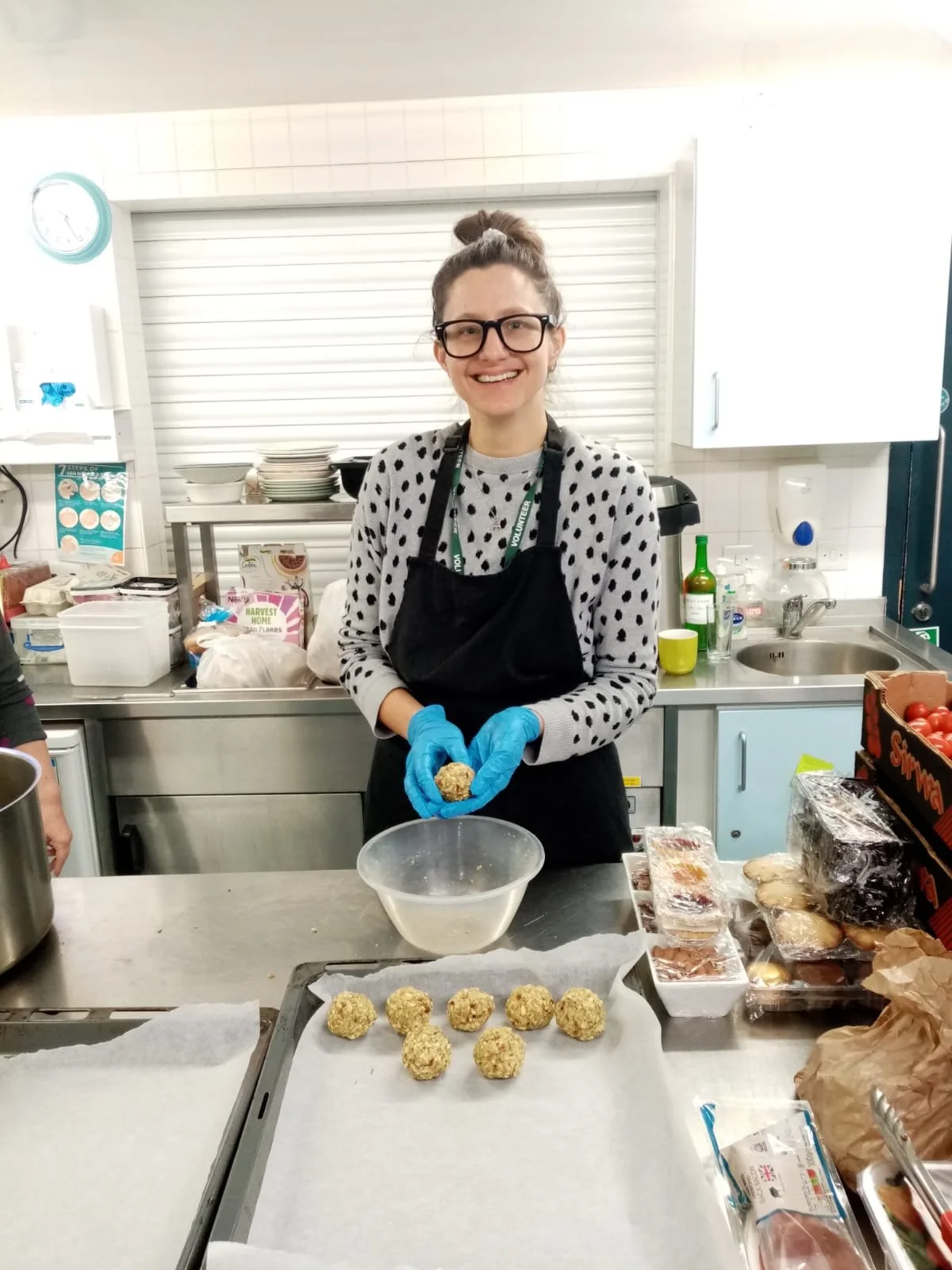 A woman wearing glasses, a gray sweater with black polka dots, a black apron, and blue gloves is smiling and holding a food mixture, standing in a kitchen surrounded by baking ingredients and supplies. In front of her on a tray, there are several round, oatmeal or cookie balls.