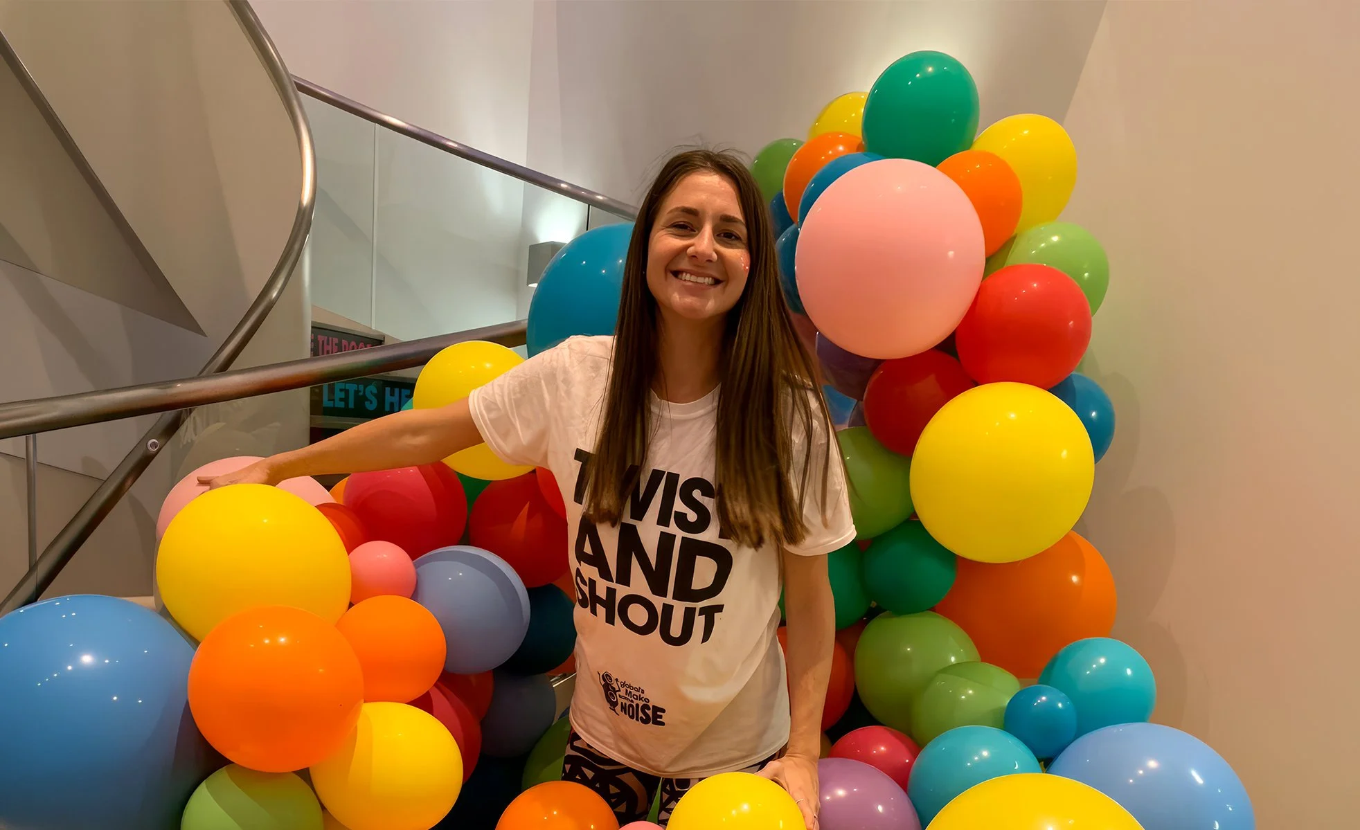 A woman smiling in front of a colorful balloon display, wearing a white T-shirt with black text. The balloons are in various colors including yellow, orange, red, blue, green, and pink, arranged in a cluster around her. She is on a staircase with a curved metal railing in an indoor setting.