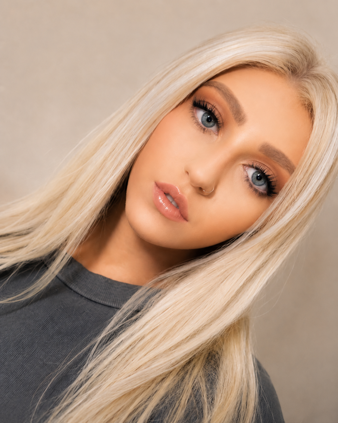 Close-up portrait of a young woman with platinum blonde hair, blue eyes, wearing makeup, a nose ring, and a dark gray top, looking at the camera against a neutral background.