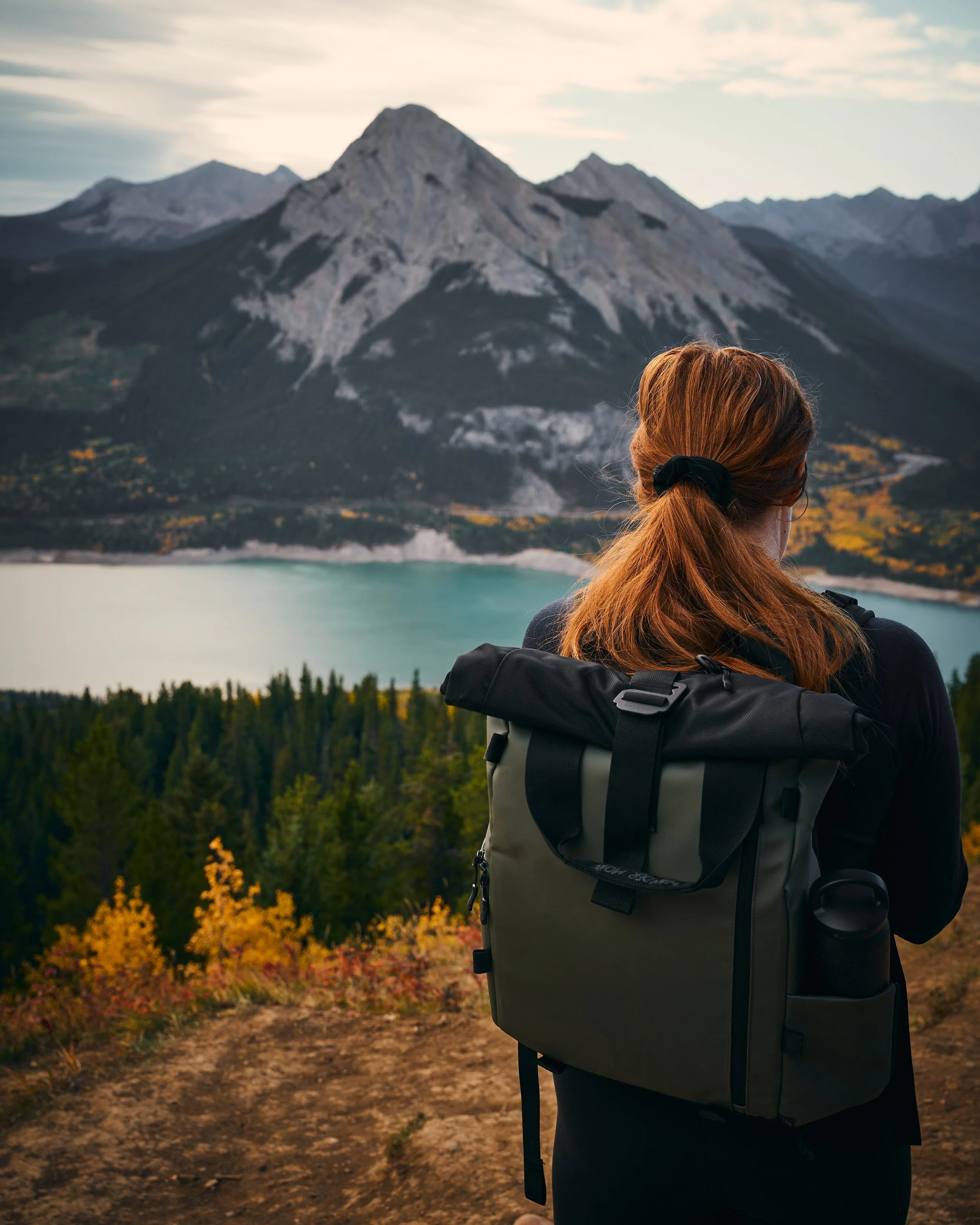 A woman with red hair tied back with a black hair tie, wearing a large backpack, looking at a mountain landscape with a lake, trees, and mountains in the background during late afternoon or early evening.