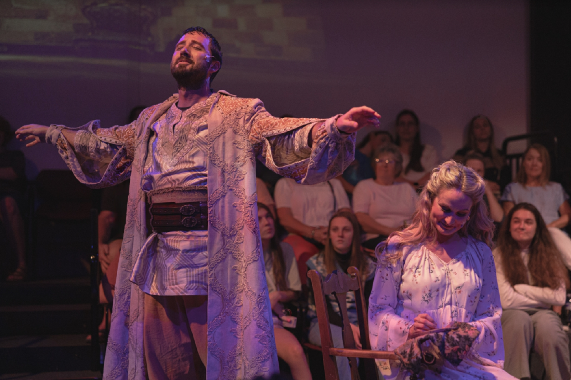 A man and woman in period costumes performing in front of an audience. The man is standing with arms outstretched, wearing a cream-colored, ornate robe, and the woman is sitting and smiling, wearing a floral dress.