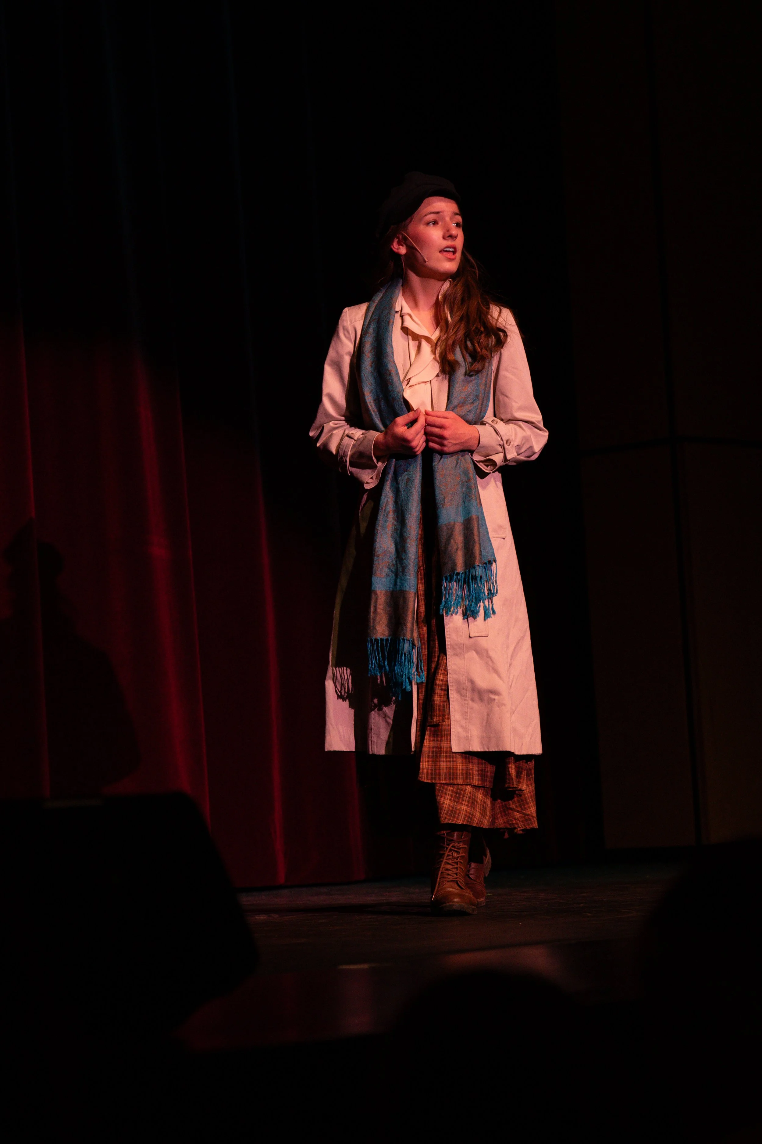 A woman is on stage, wearing a long beige coat, brown plaid pants, brown boots, a light-colored shirt, a blue scarf, and a black beret. She appears to be singing with a facial expression of concern or emotion, against a dark background with red stage curtains.